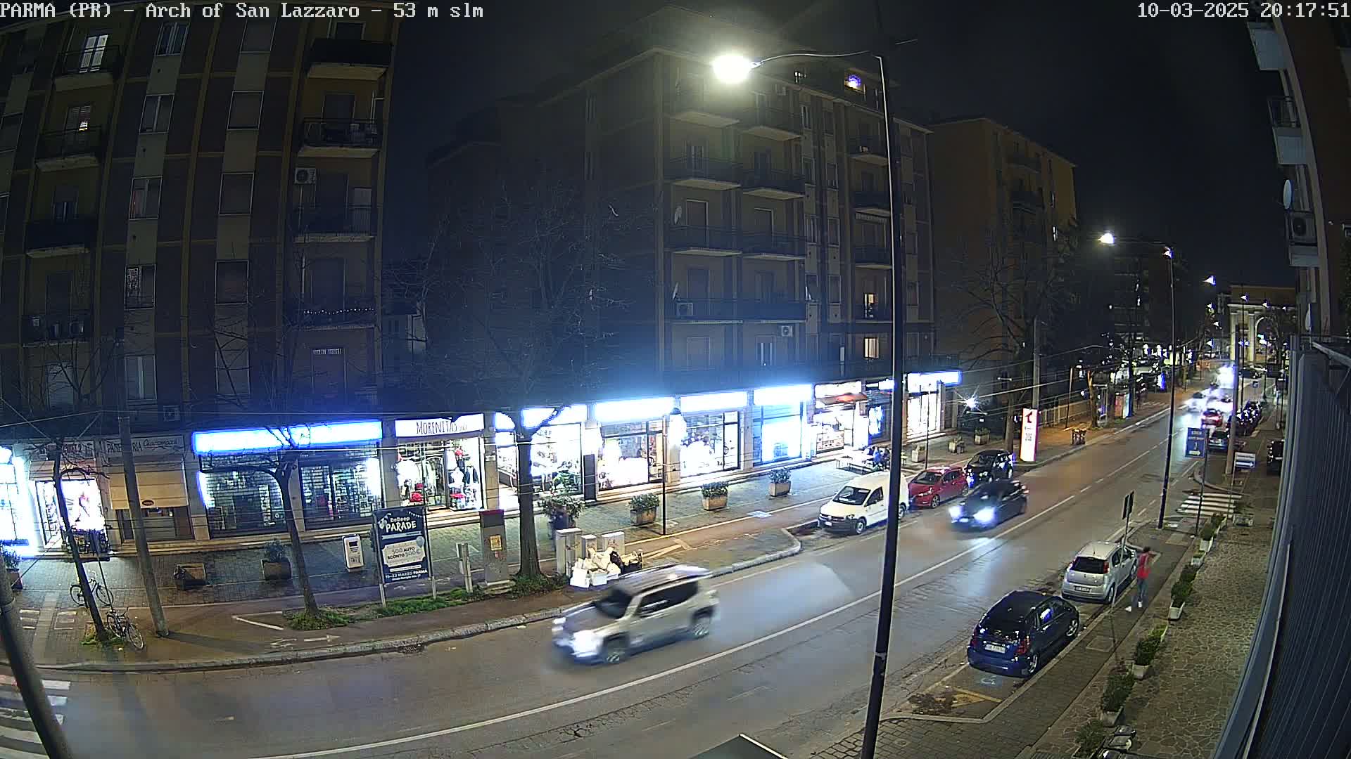 A nighttime street scene shows several cars driving on a two-lane road past shops with illuminated signs, under a dark sky.