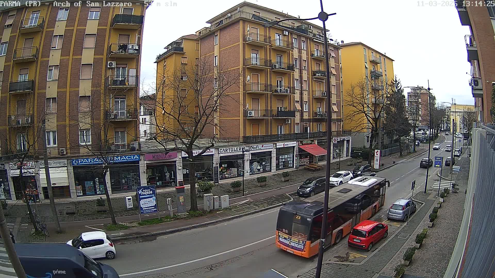 A city street scene on an overcast day shows a multi-story yellow building with shops on the ground floor, a bus and several cars on the street, and bare trees lining the sidewalk.