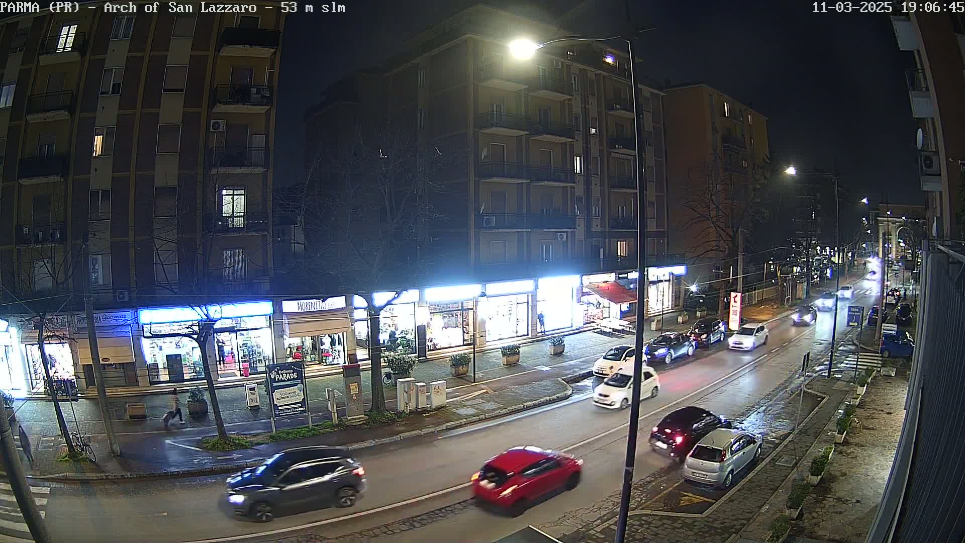 A nighttime street scene shows several cars driving on a wet road, past shops and apartment buildings under streetlights.
