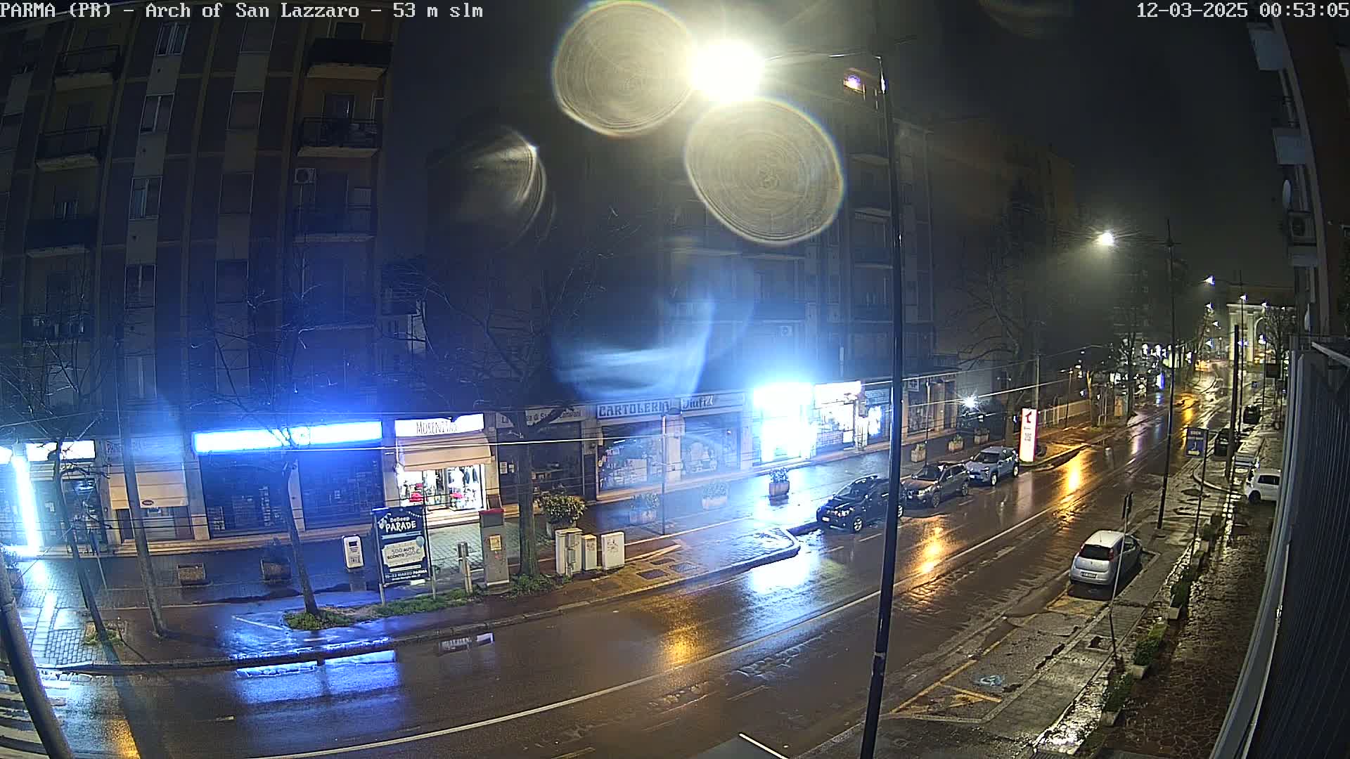 A rain-slicked street at night, showing several cars parked and driving, under bright streetlights illuminating buildings and storefronts.
