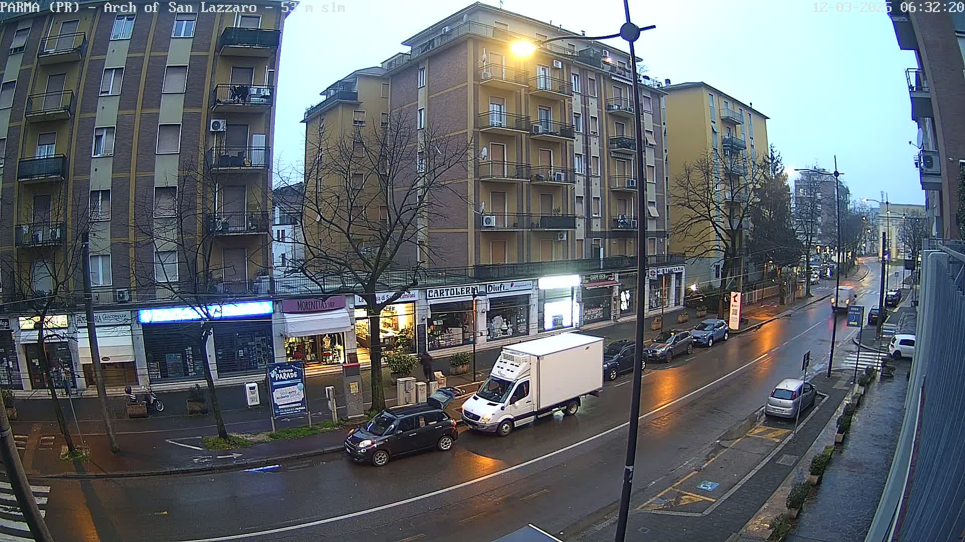 A wet city street at dawn, lined with multi-story buildings and shops, with several cars parked and driving on the road.