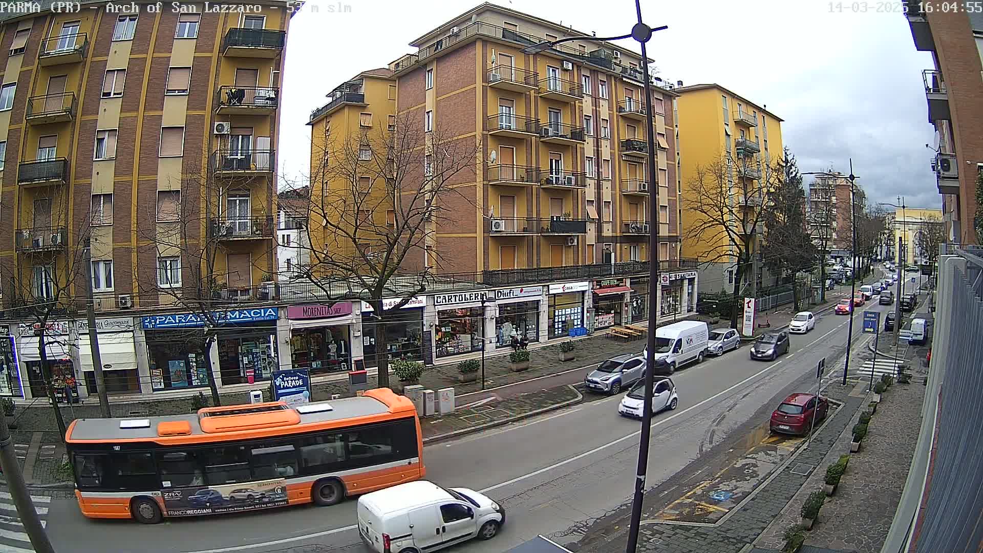 An orange bus and several cars are driving down a city street lined with multi-story buildings and shops under a cloudy sky.