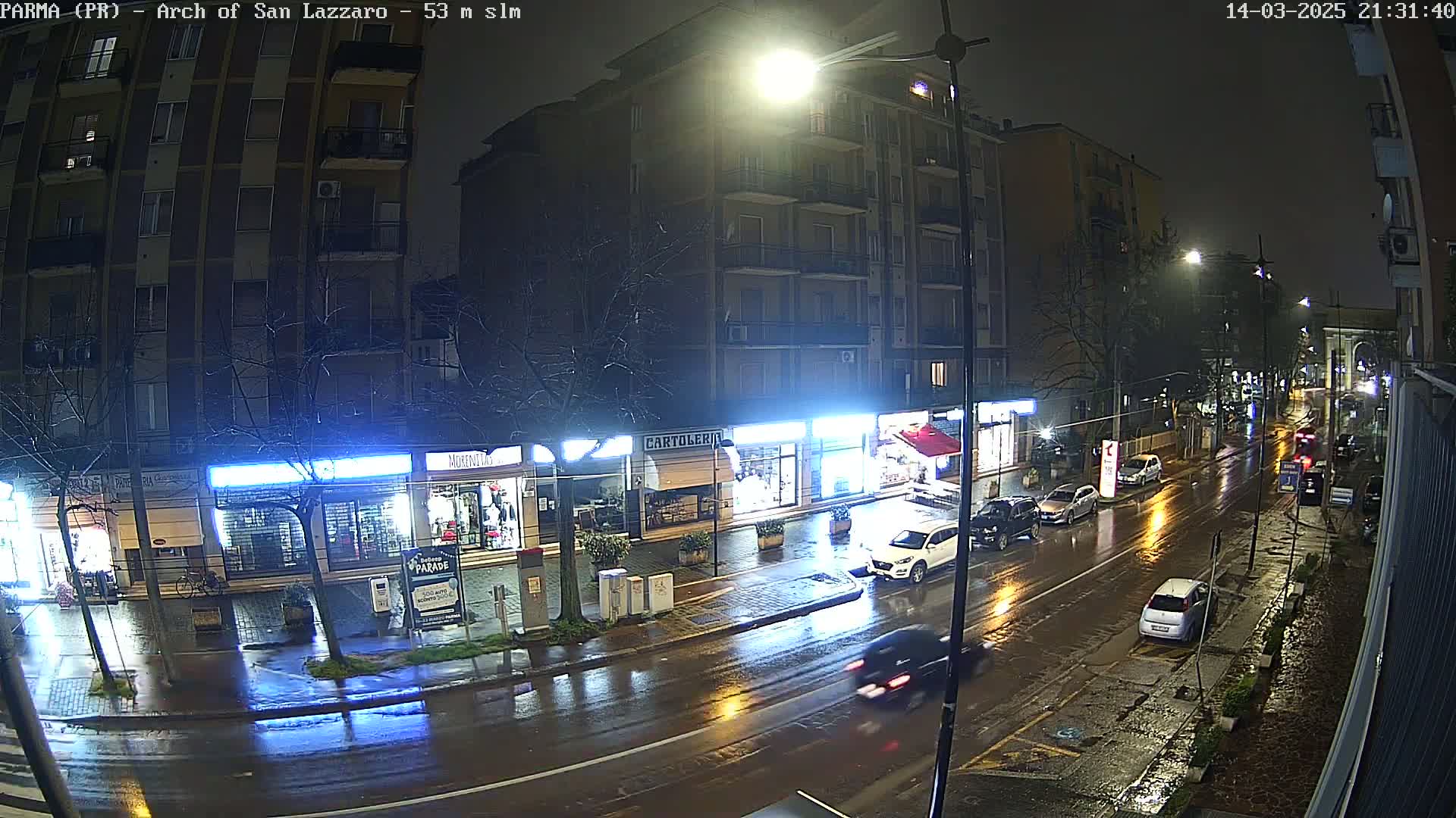 A rain-slicked street at night, lined with shops and buildings, shows several cars driving under streetlights.