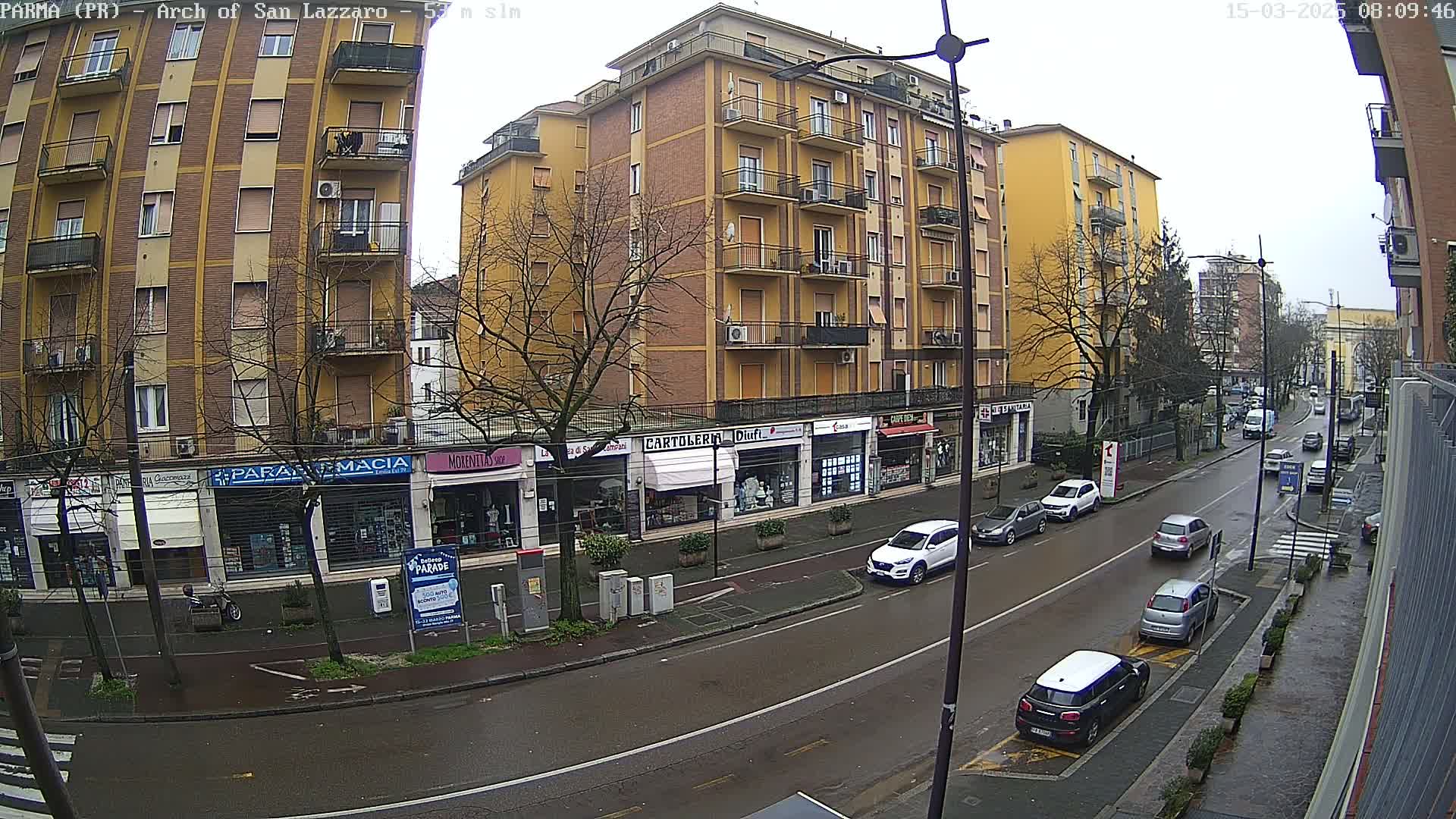A street scene on an overcast day shows a multi-story building with shops on the ground floor, cars parked along the curb, and several vehicles traveling down a wet road.