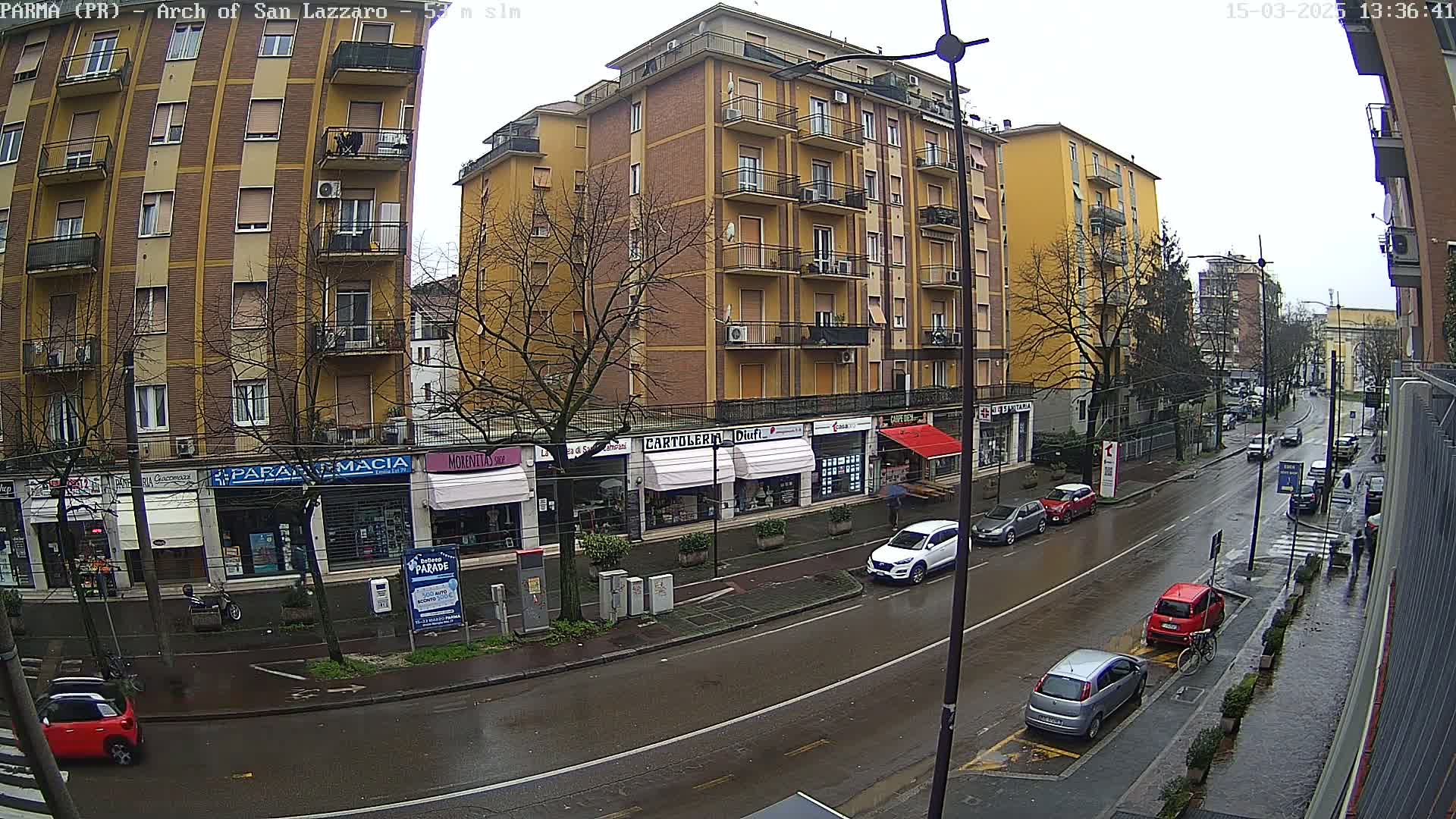 A wet city street scene shows several cars parked and driving alongside multi-story buildings on a rainy day.