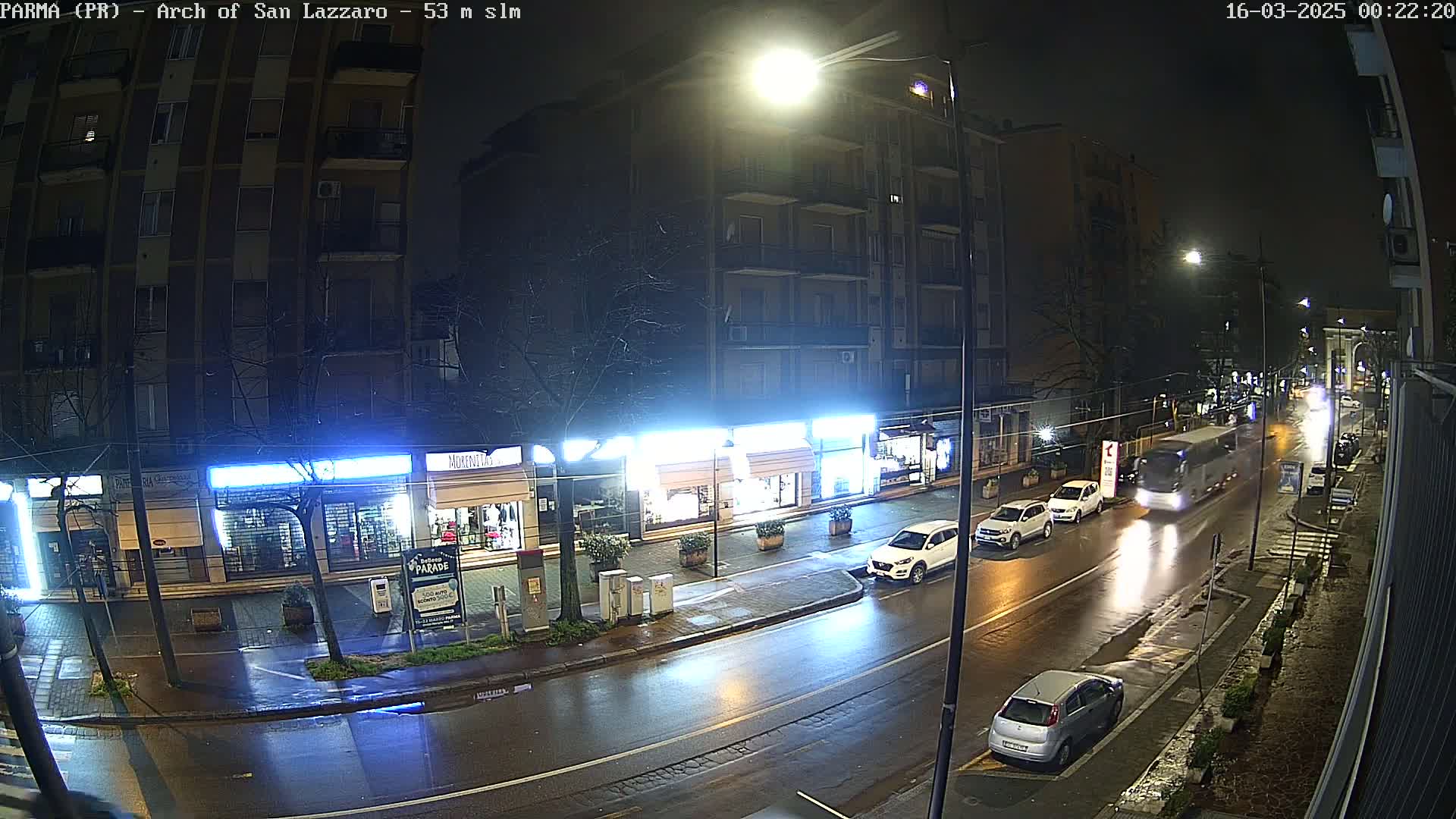 A wet city street at night, illuminated by streetlights, shows several cars parked along the side of the road and a bus driving down the center, with buildings lining the street.
