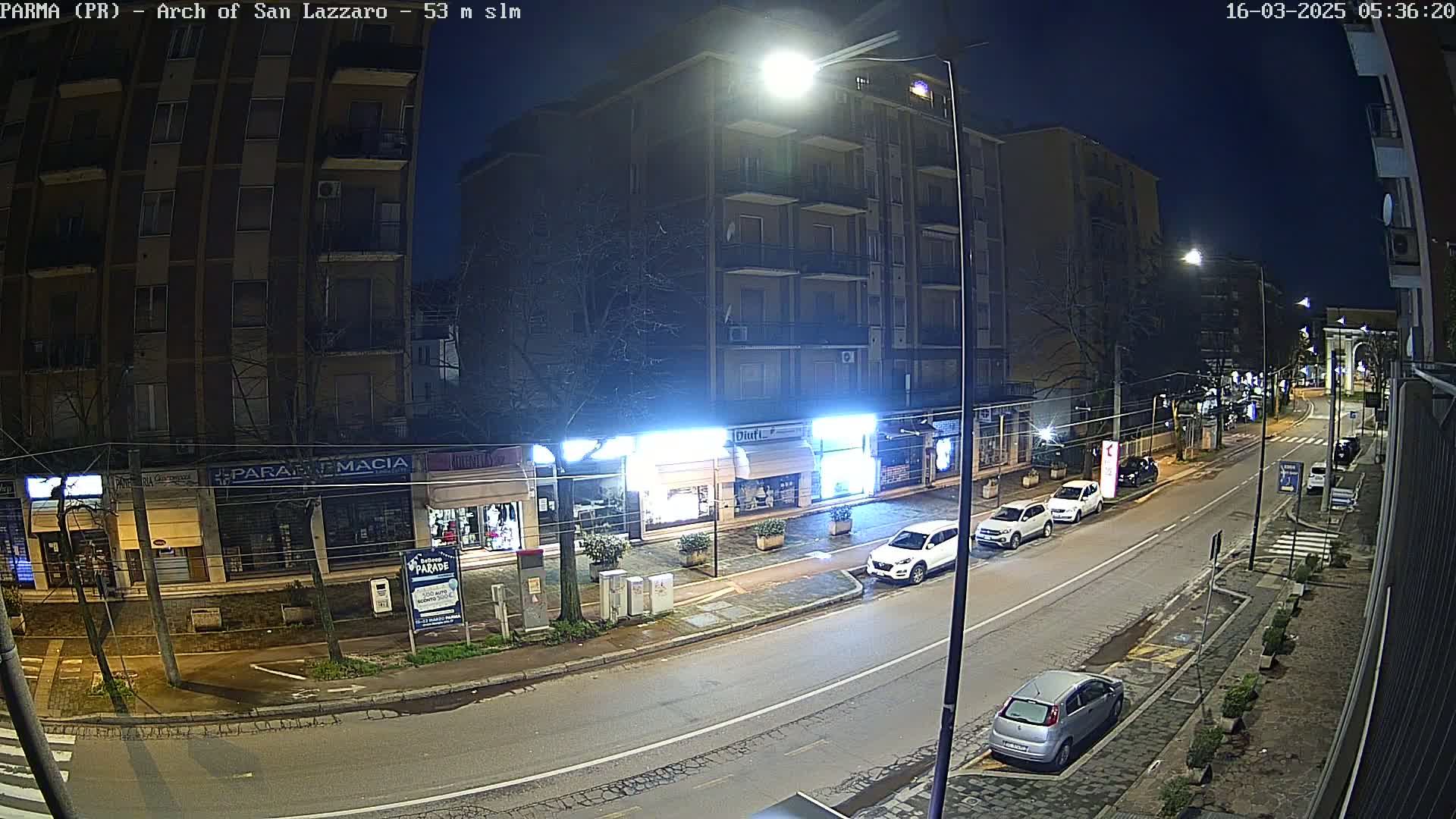 A nighttime street scene shows several parked cars in front of shops and apartment buildings under bright streetlights.