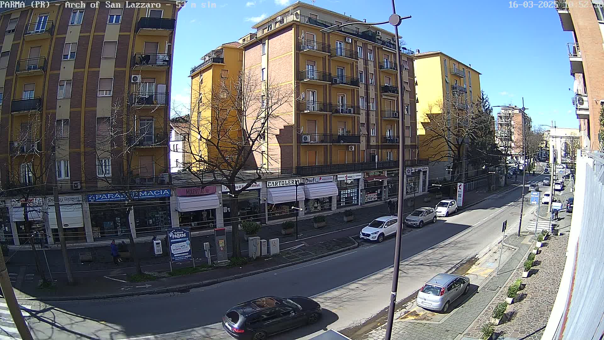 A sunny day on a city street, showing a multi-story yellow building with shops on the ground floor and cars parked along the roadside.