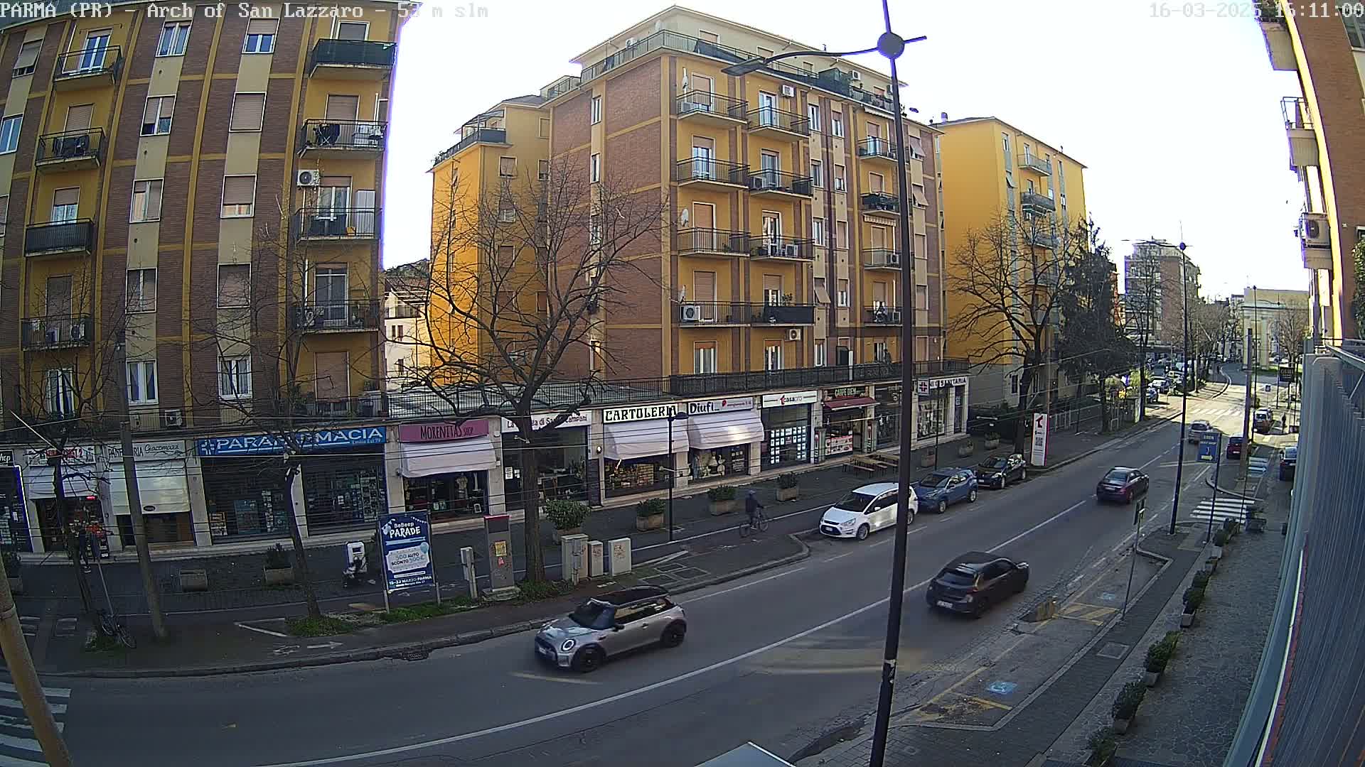 A sunny city street scene shows a multi-story yellow building with shops on the ground floor, several cars parked and driving on the street, and bare trees lining the sidewalk.