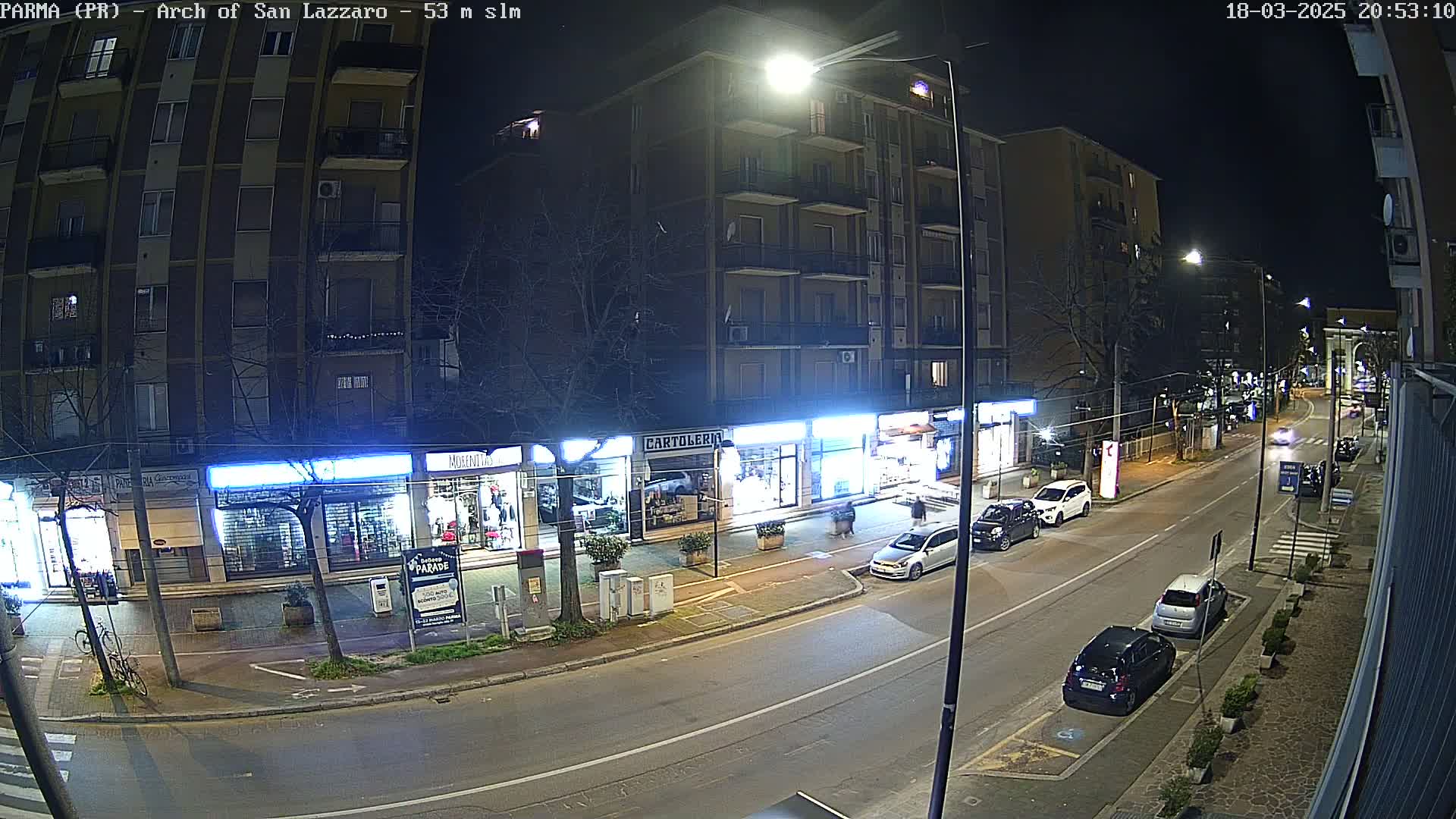 A nighttime street scene shows several parked cars along a city street lined with storefronts under bright streetlights.