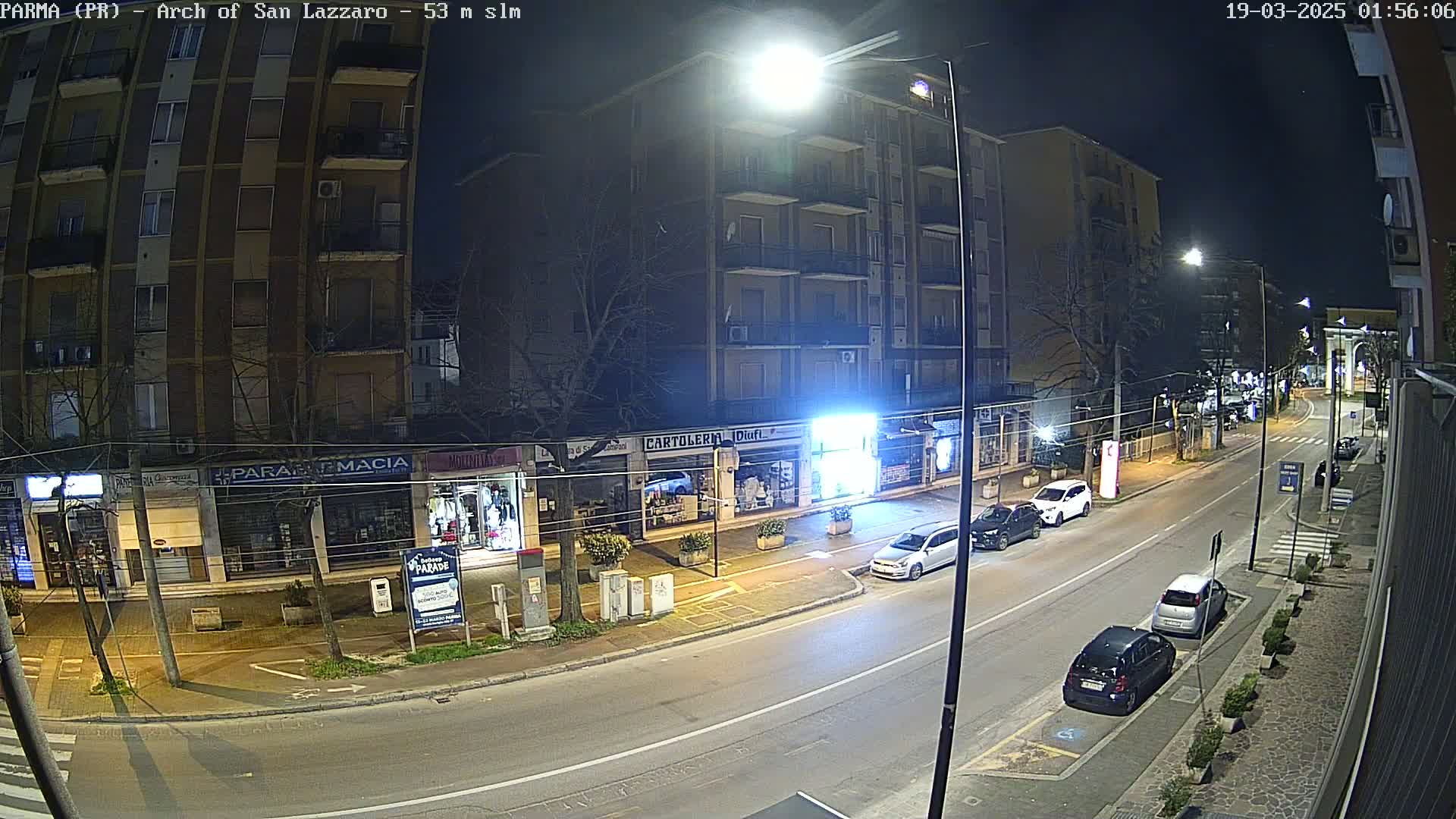 A nighttime street scene shows several parked cars along a multi-lane road lined with shops and apartment buildings under streetlights.