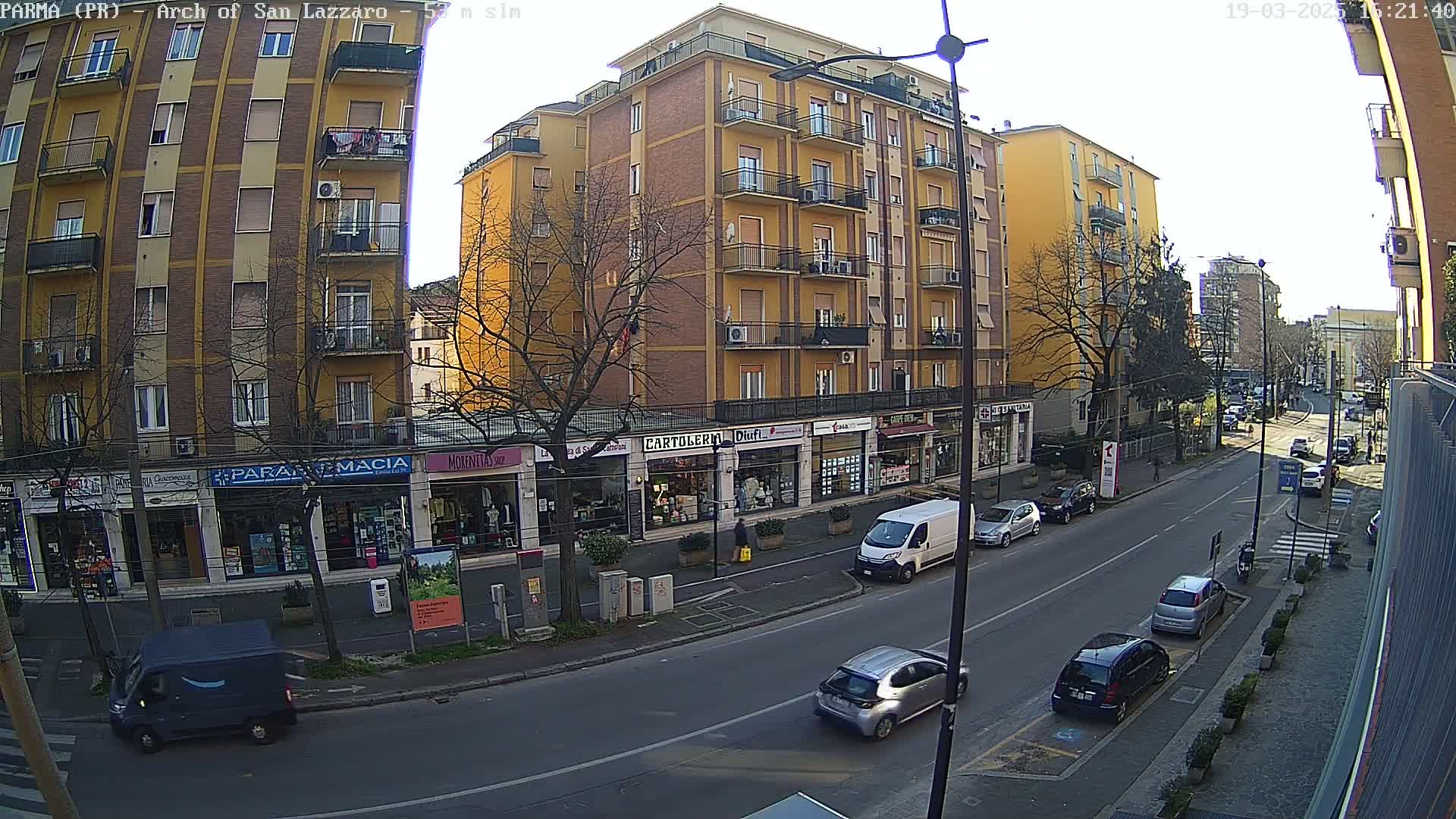 A sunny street scene shows several cars parked and driving along a road lined with multi-story buildings and shops.