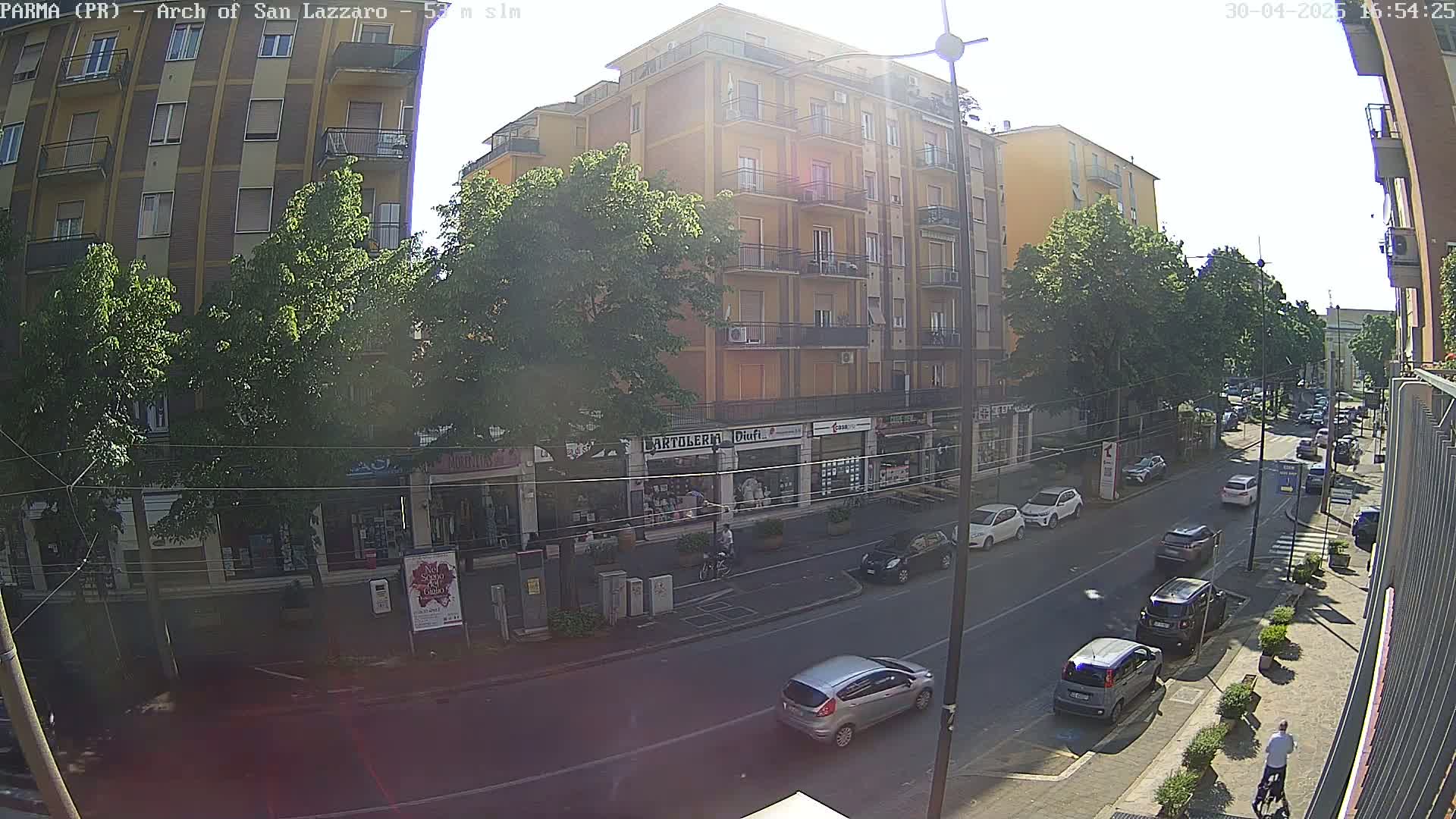 A sunny street scene shows a multi-story building with shops on the ground floor, several cars parked and driving along a road lined with trees, and a few pedestrians.