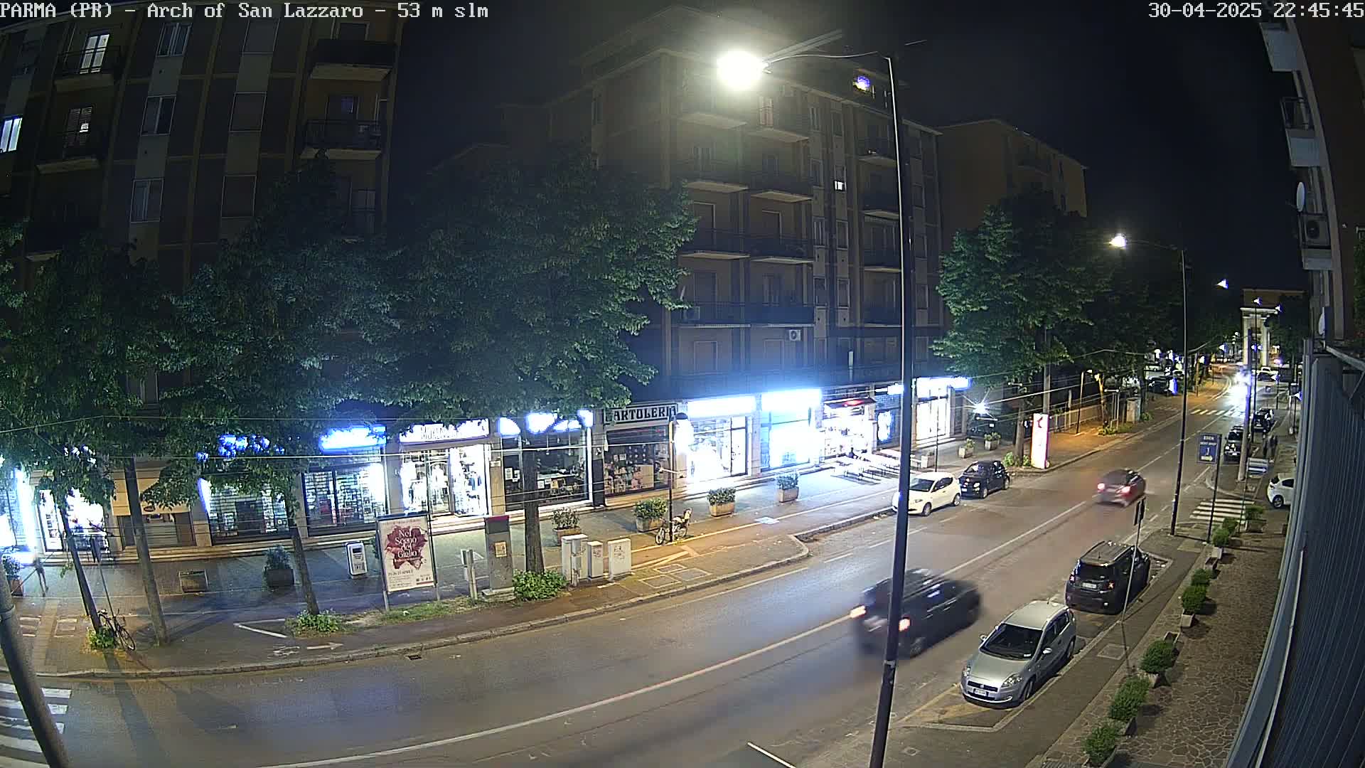 A nighttime street scene shows a row of shops and apartment buildings under streetlights, with several cars parked and driving on the road.