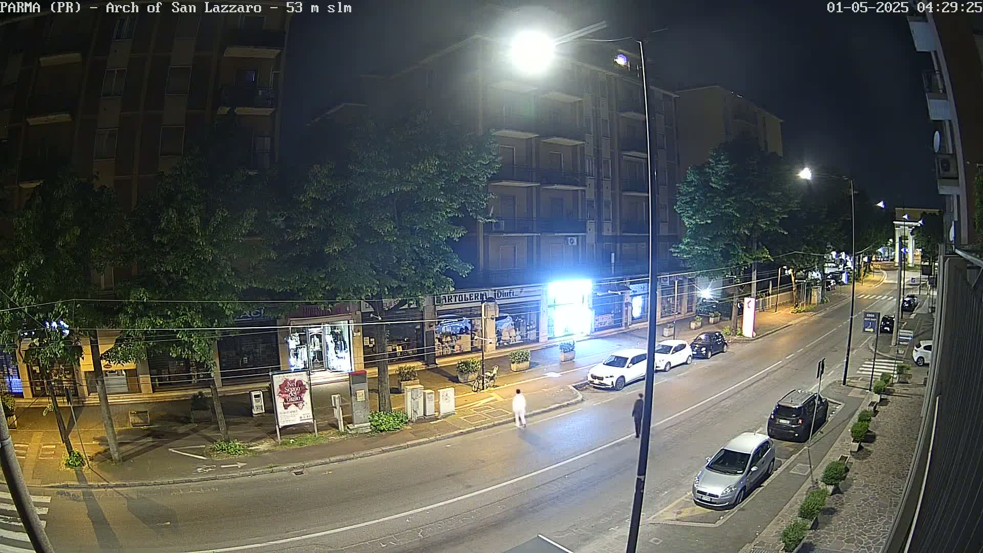 A nighttime street scene shows several parked cars and two pedestrians on a mostly empty road lined with shops and trees under bright streetlights.