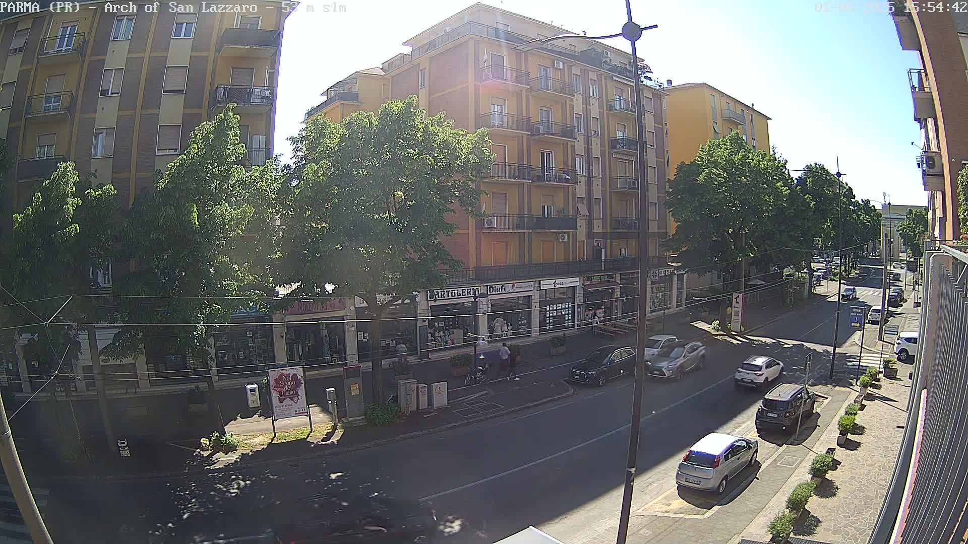 A sunny city street scene shows several parked and moving cars in front of multi-story buildings with shops on the ground floor, lined with leafy trees.