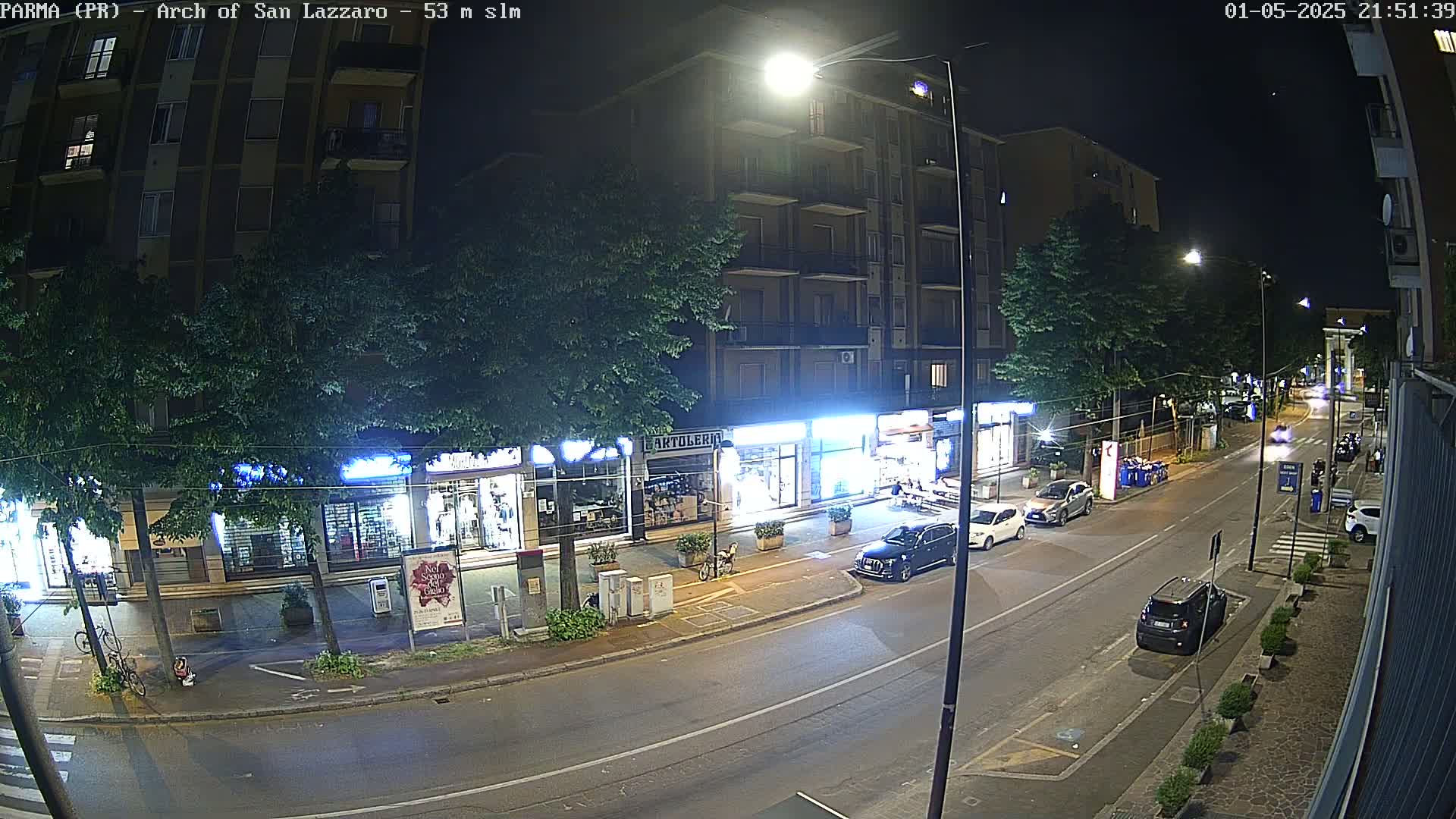 A nighttime street scene shows a row of shops and a multi-story building illuminated by streetlights, with several cars parked along the road and a few driving by.