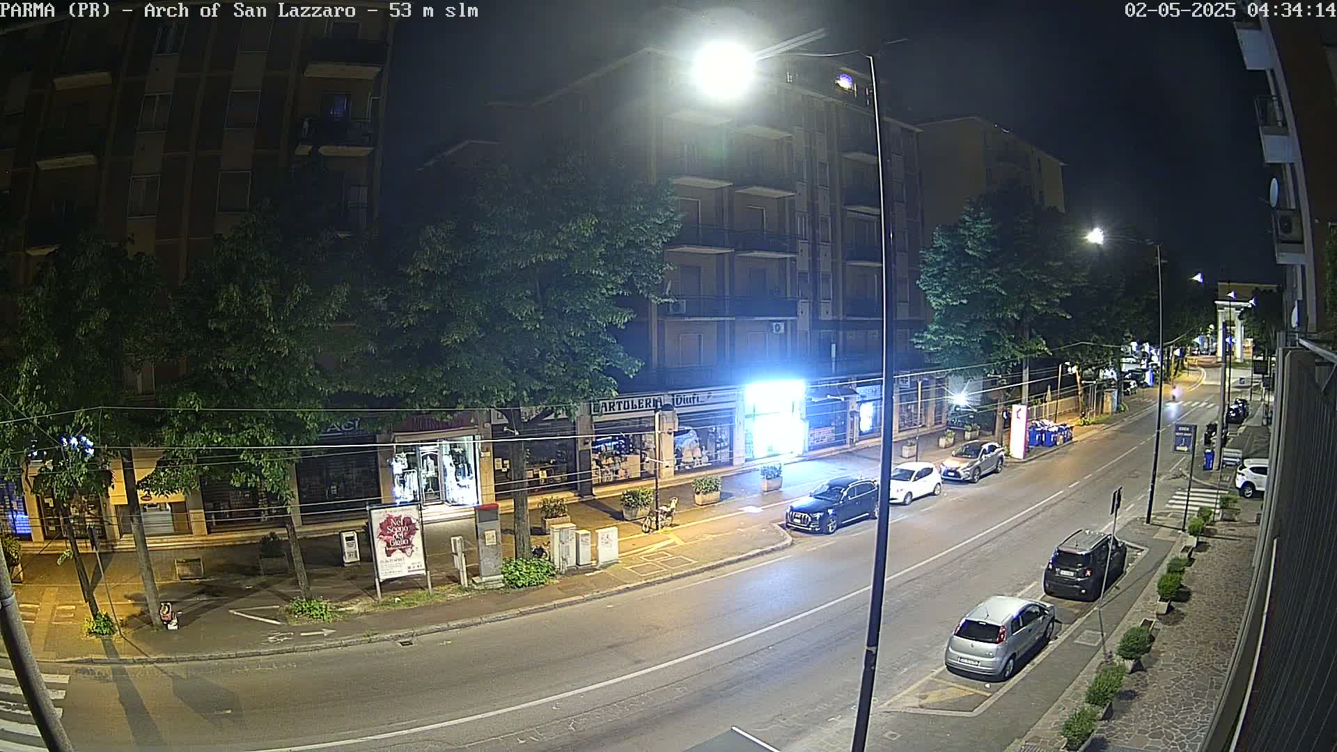 A nighttime street scene shows a multi-story building with storefronts, several parked cars, and a few streetlights illuminating the road.