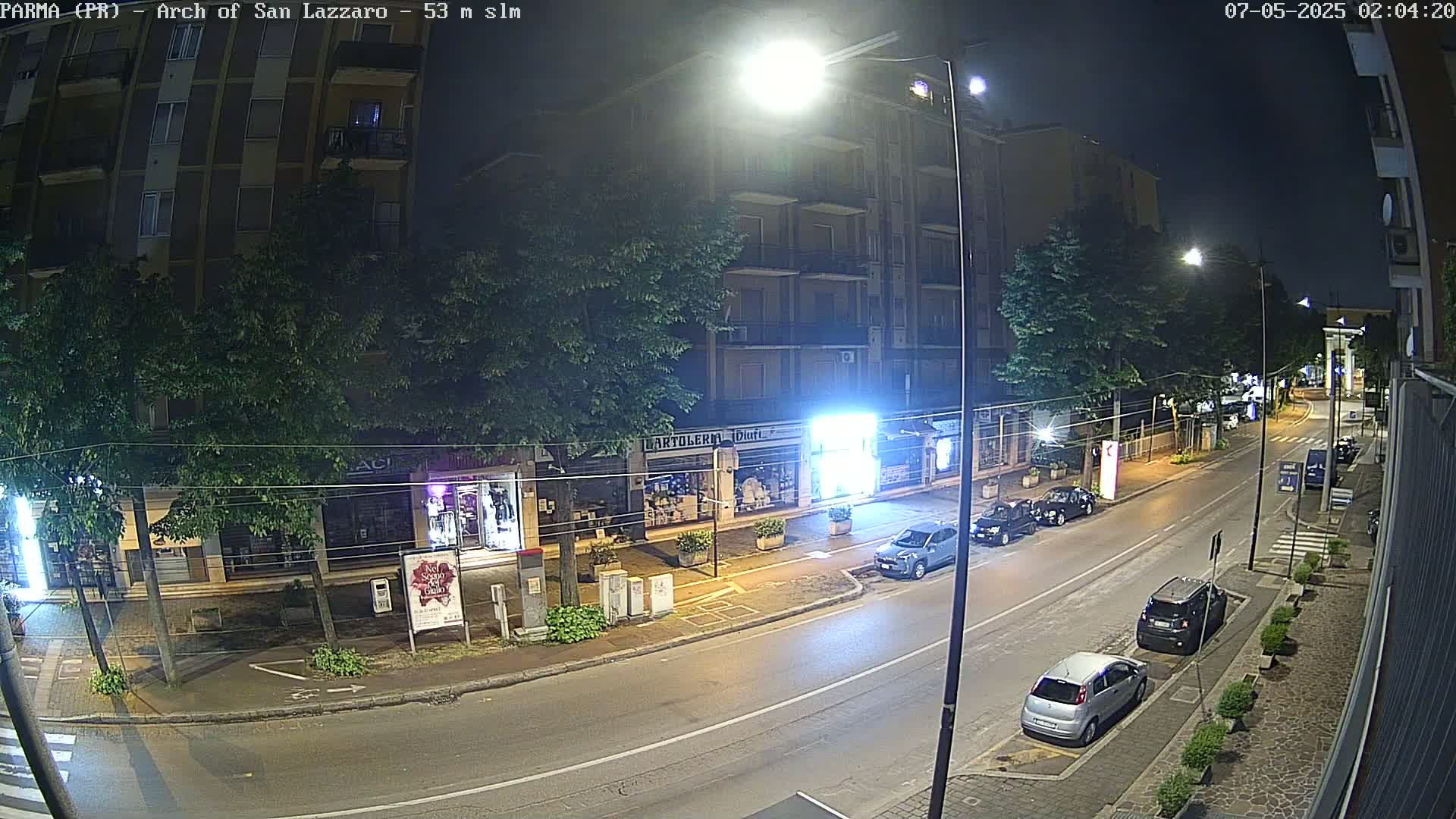 A nighttime street scene shows a multi-story building with street-level shops and several parked cars under bright streetlights.
