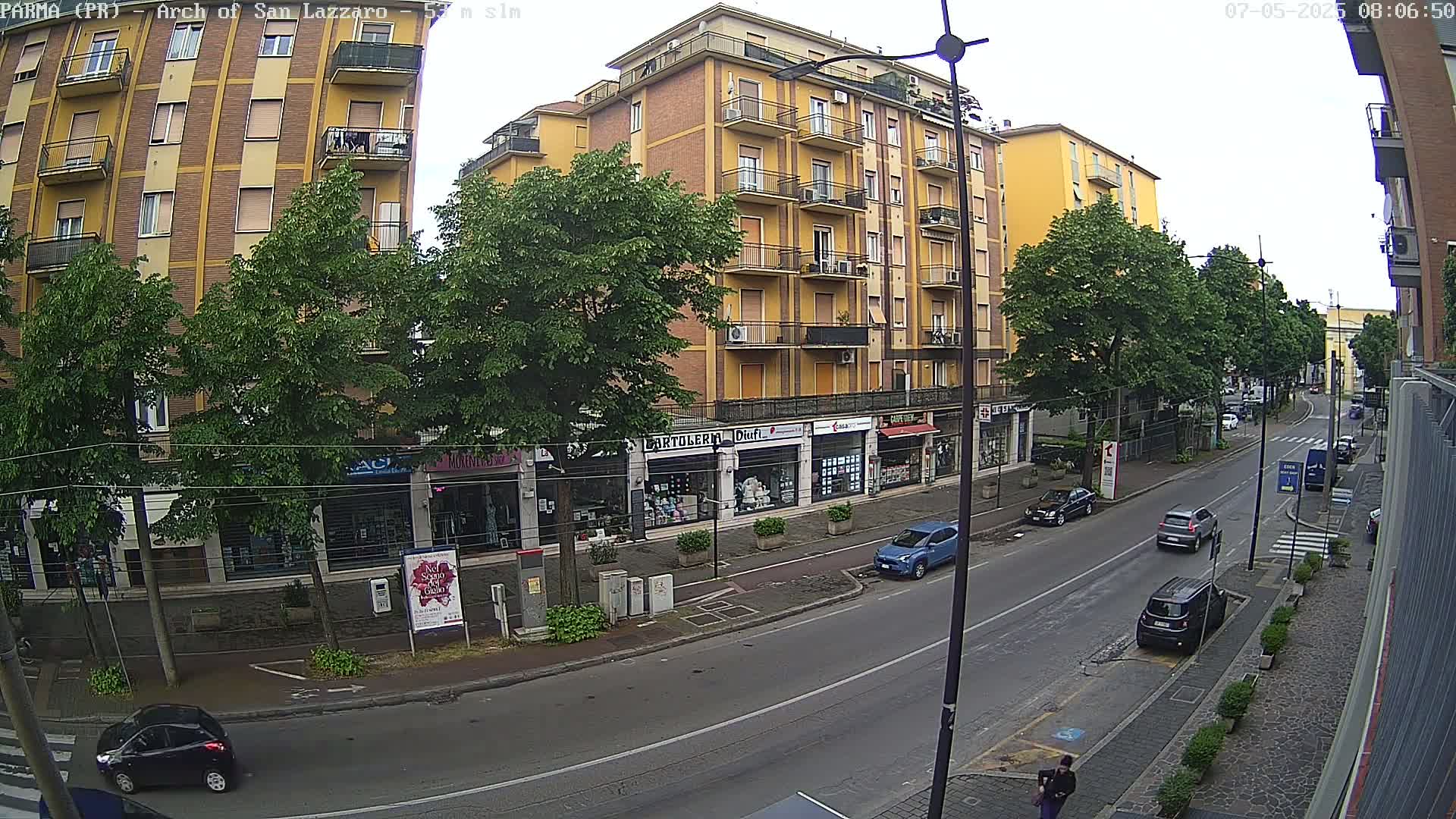 A mostly sunny day shows a street scene with several cars parked and driving past a multi-story building with shops on the ground floor, flanked by trees.