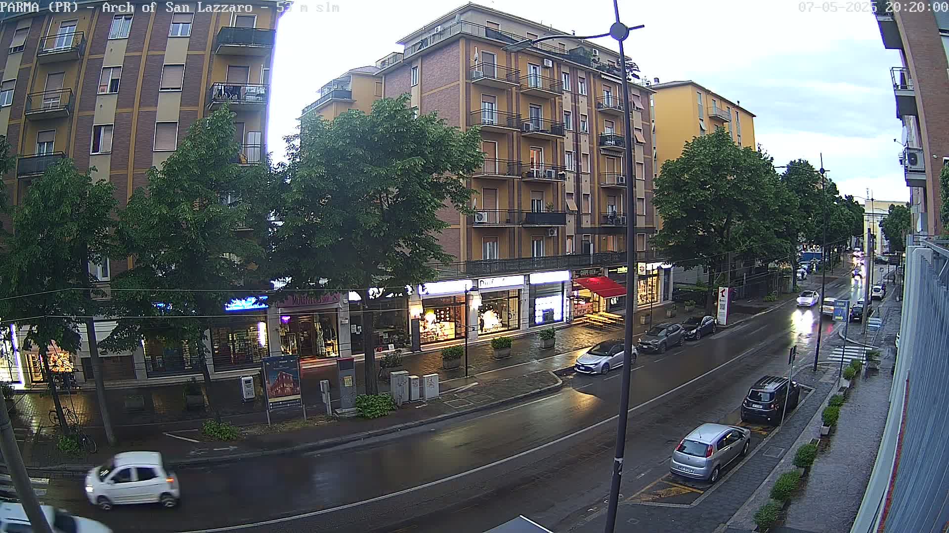 A wet street lined with shops and apartment buildings under an overcast sky, with several cars parked and driving on the road.