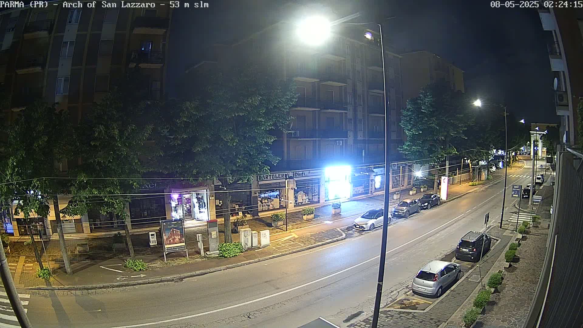A nighttime street scene shows a mostly empty road with parked cars alongside buildings under streetlights.