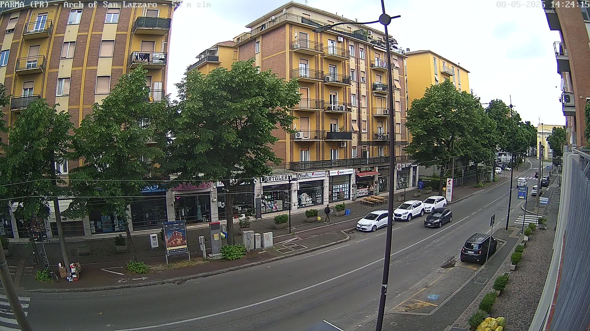 A partly sunny street scene shows a multi-story yellow building with shops on the ground floor, several parked cars, and leafy green trees lining the street.