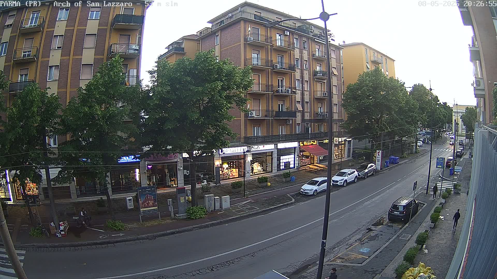 A street scene under an overcast sky shows several multi-story buildings, shops with outdoor displays, parked cars, and a few pedestrians.
