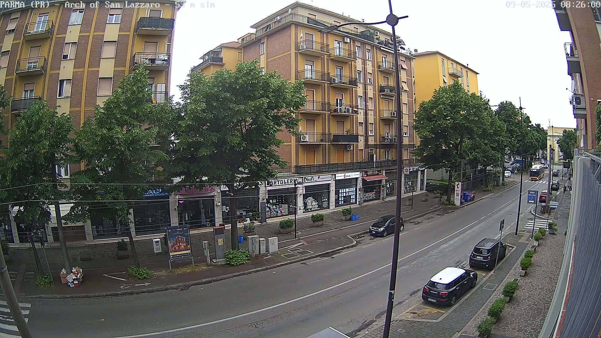 A street scene on an overcast day shows a multi-story yellow building with shops on the ground floor, several cars parked along the street, and lush green trees lining the sidewalk.