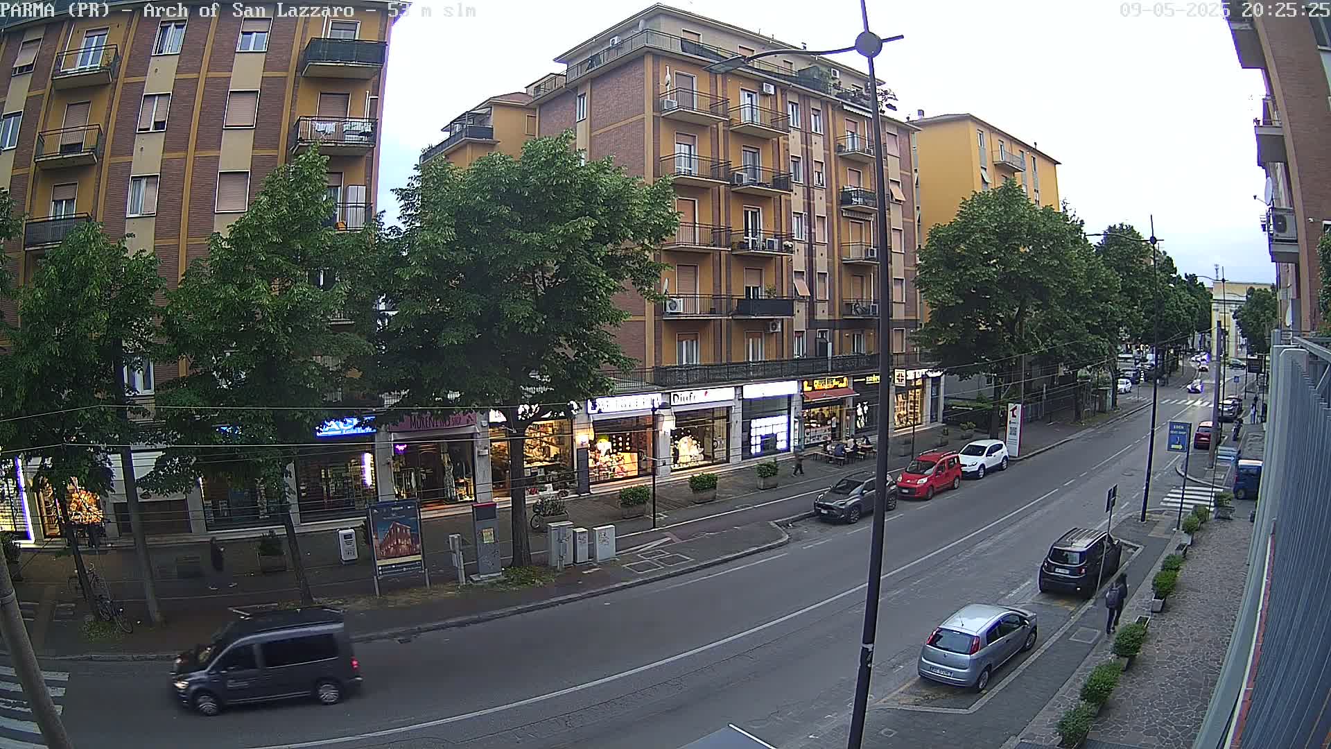 A city street scene at dusk shows a multi-story building with shops on the ground floor, several cars parked along the street, and pedestrians walking on the sidewalk.