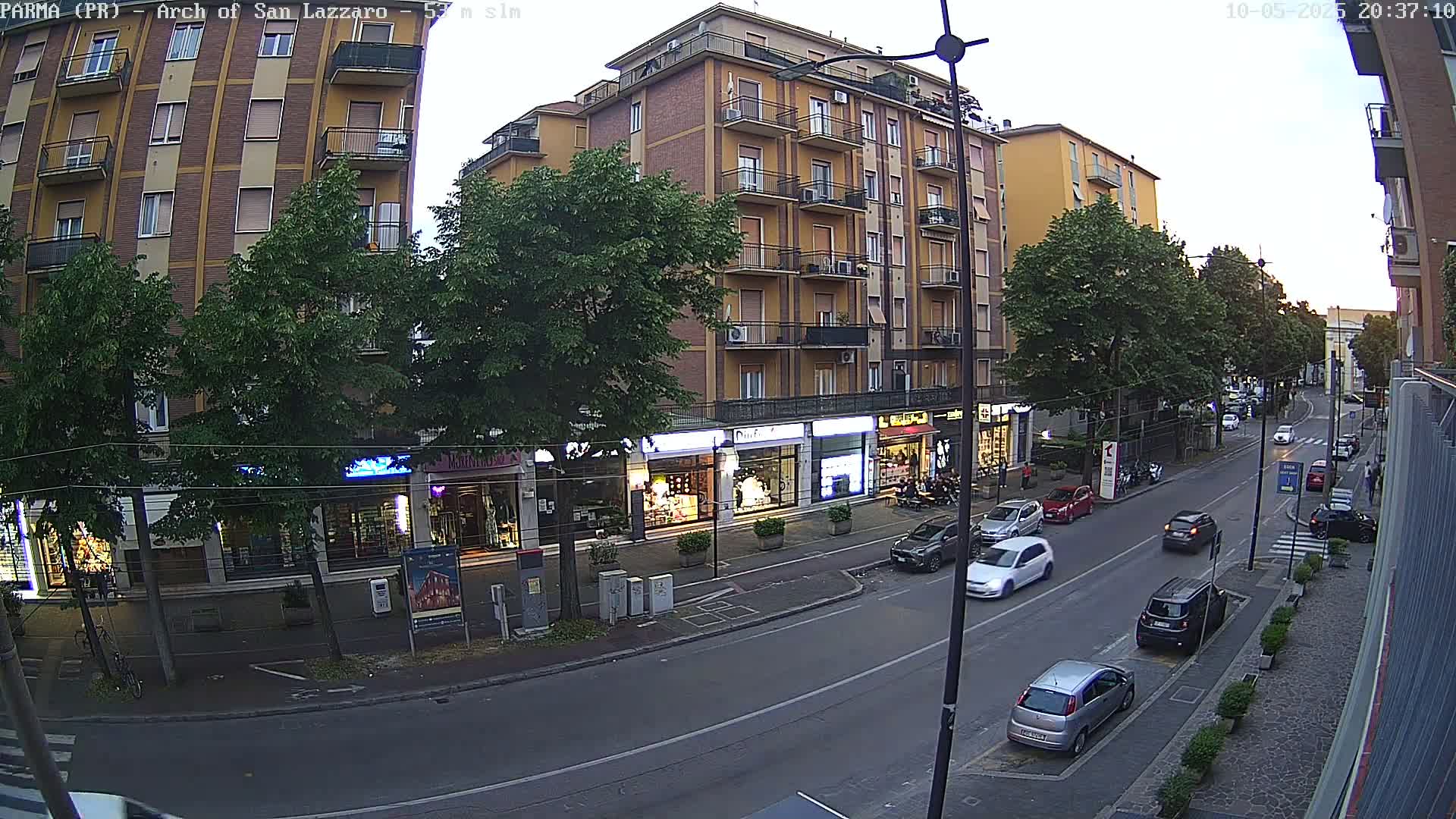 A street lined with multi-story buildings and shops, under a twilight sky, has several cars parked and driving on it.