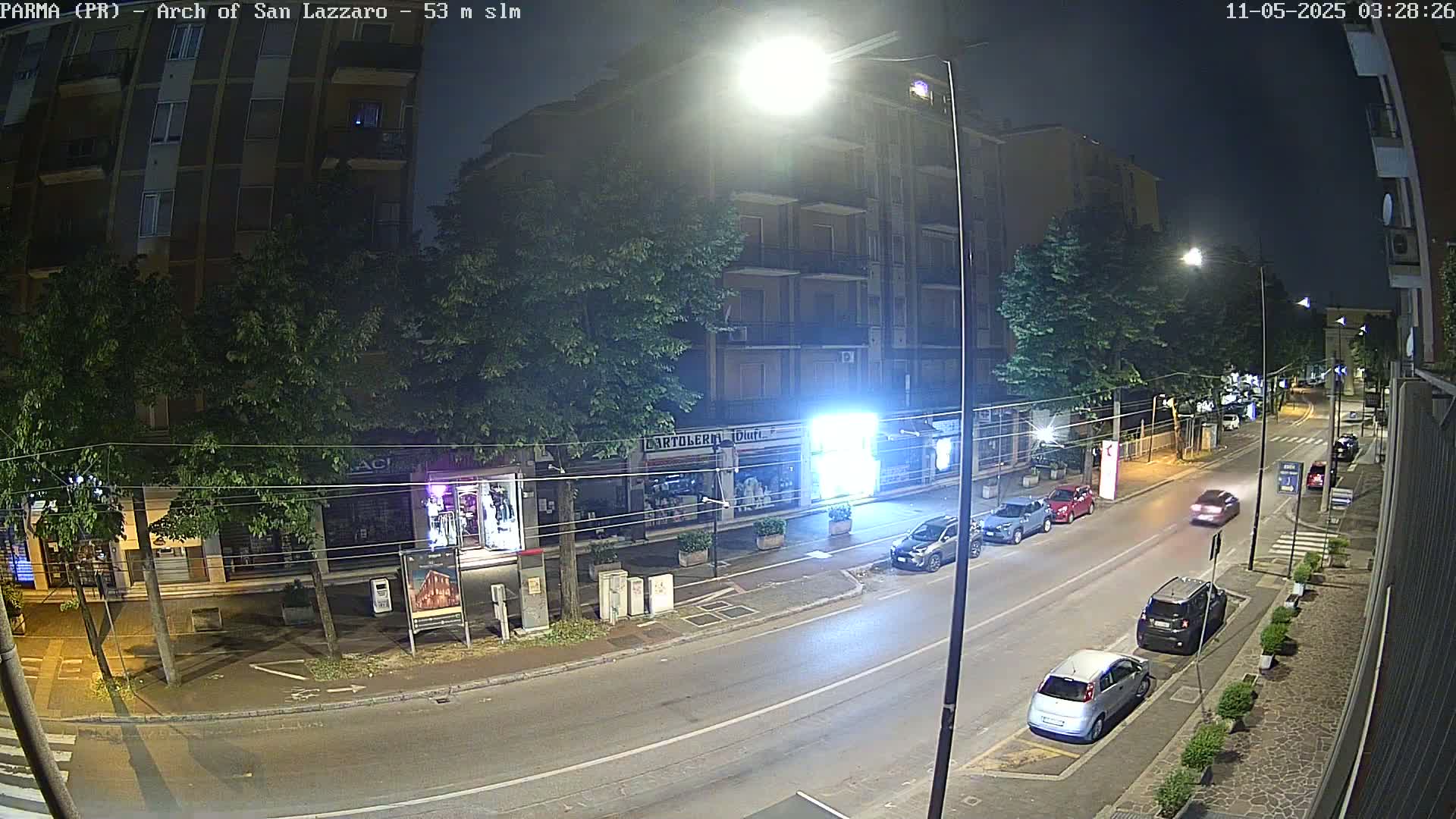 A nighttime street scene shows a multi-story building and several parked cars, under streetlights on a mostly clear night.