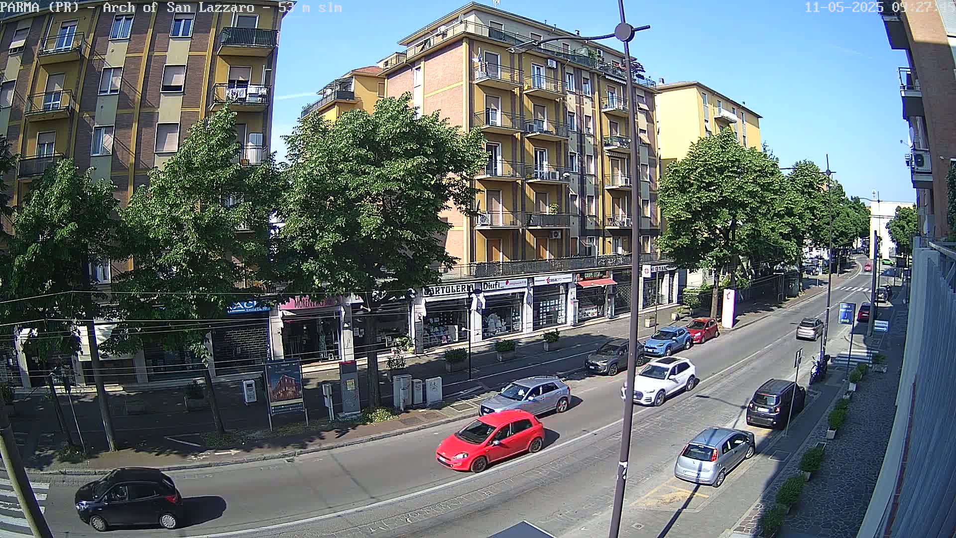 A sunny day reveals a city street scene with several cars parked and driving past multi-story buildings and shops, lined with leafy trees.