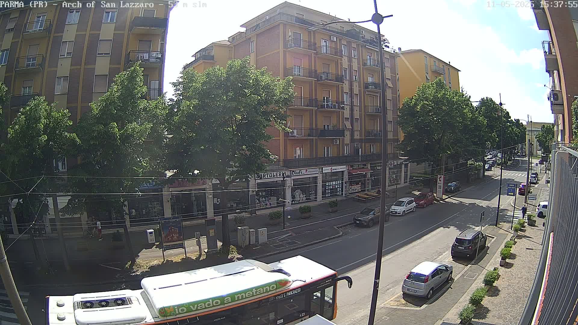 A sunny street scene shows a row of buildings with shops on the ground floor, several cars parked along the side of the road, and a bus in the foreground.