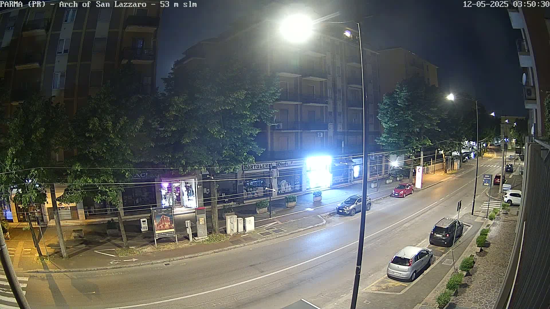 A nighttime street scene shows a mostly empty road with parked cars, streetlights, and several storefronts along the side of a multi-story building.