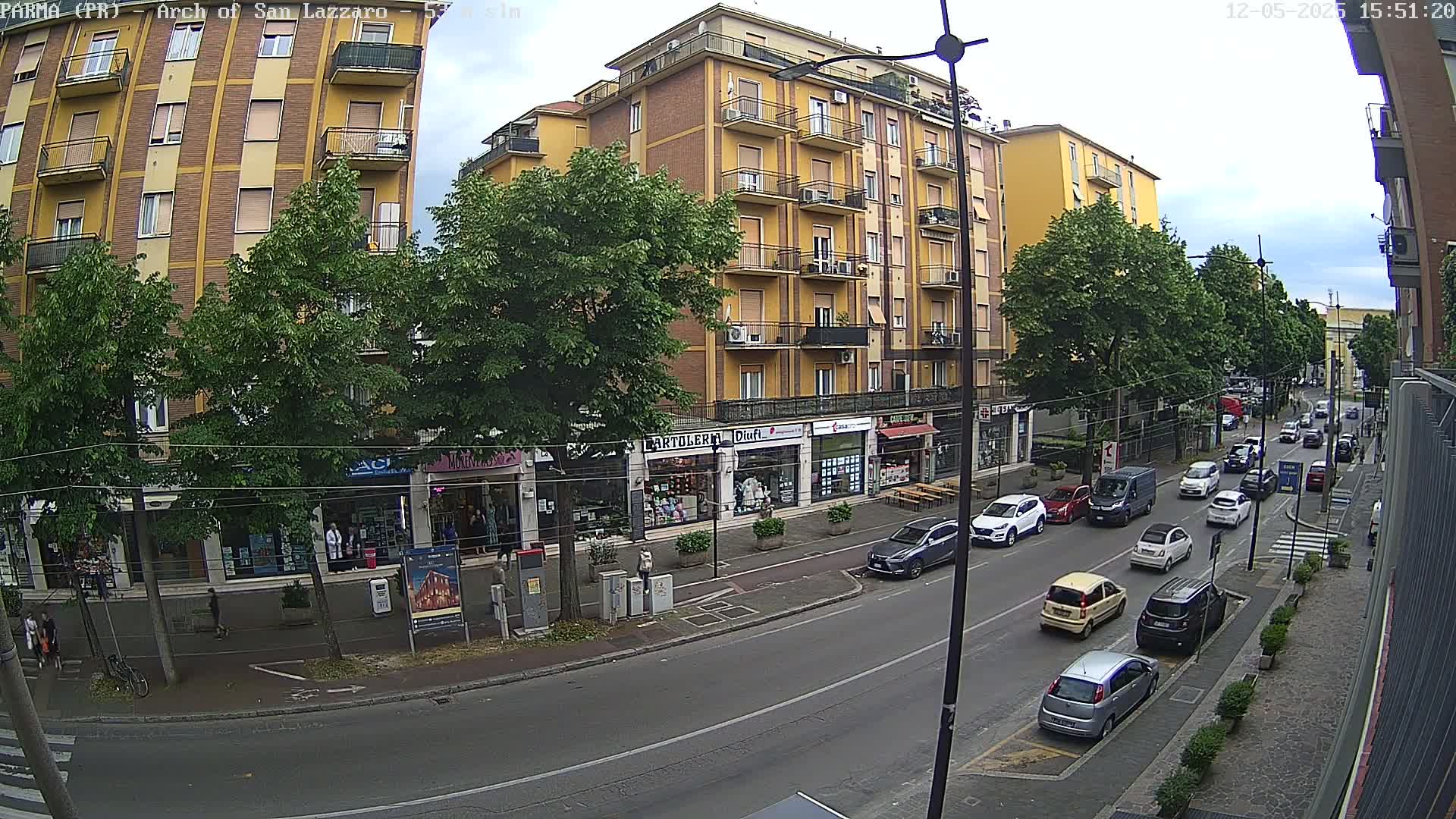 A street scene on a partly cloudy day shows a row of multi-story buildings with shops on the ground floor, and cars parked and driving on the street in front.