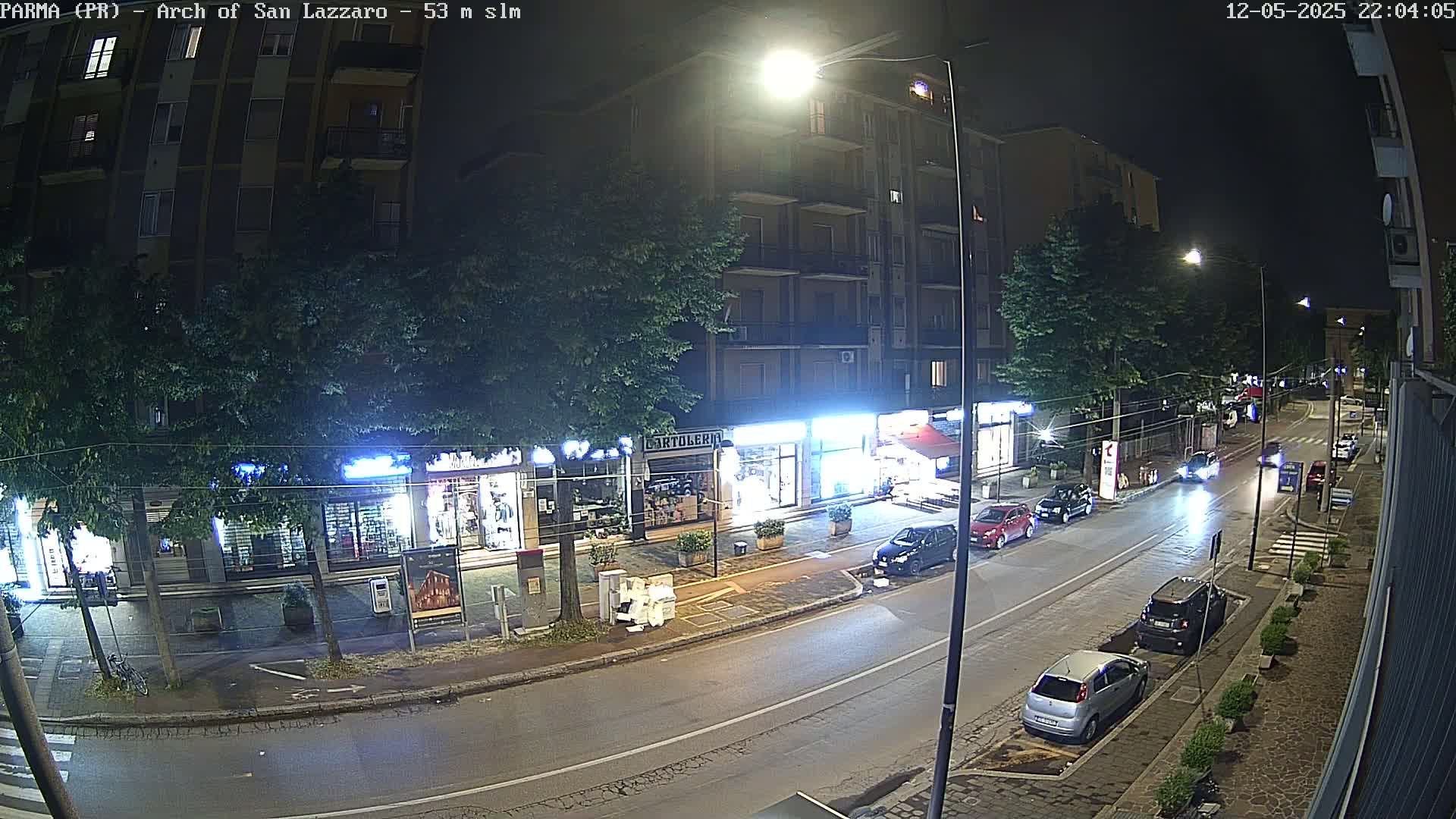 A nighttime street scene shows a row of shops and buildings with cars parked along the side of the road, under streetlights.