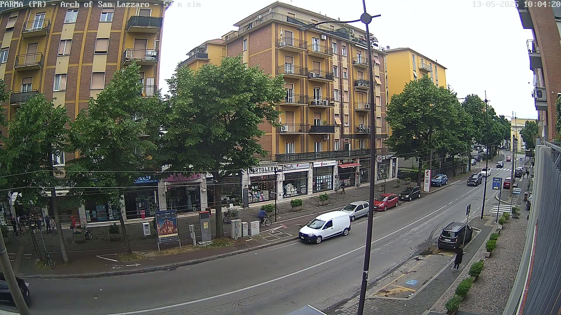 A street scene on an overcast day shows a multi-story yellow building with shops on the ground floor, several cars parked and driving along the street, and people walking on the sidewalk.