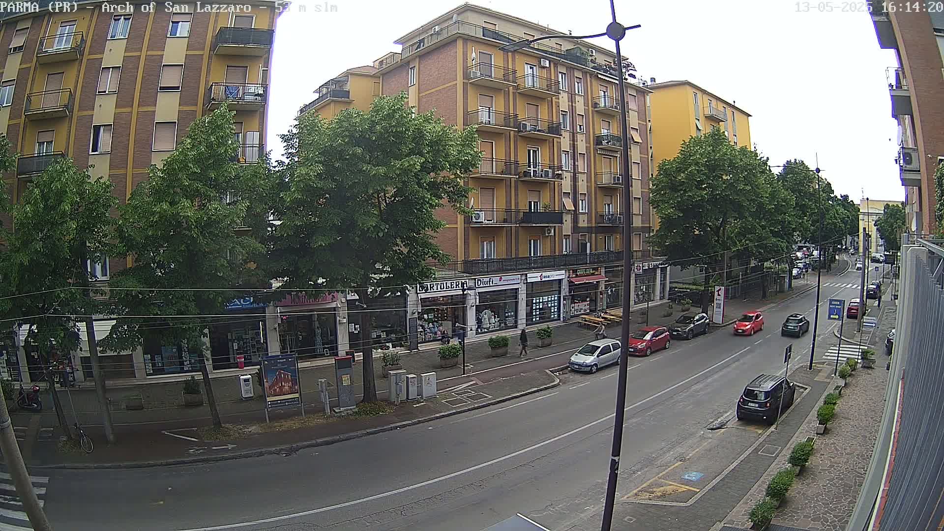 A city street scene on an overcast day shows a multi-story building with shops on the ground floor, lined with green trees, and several cars parked along the street.