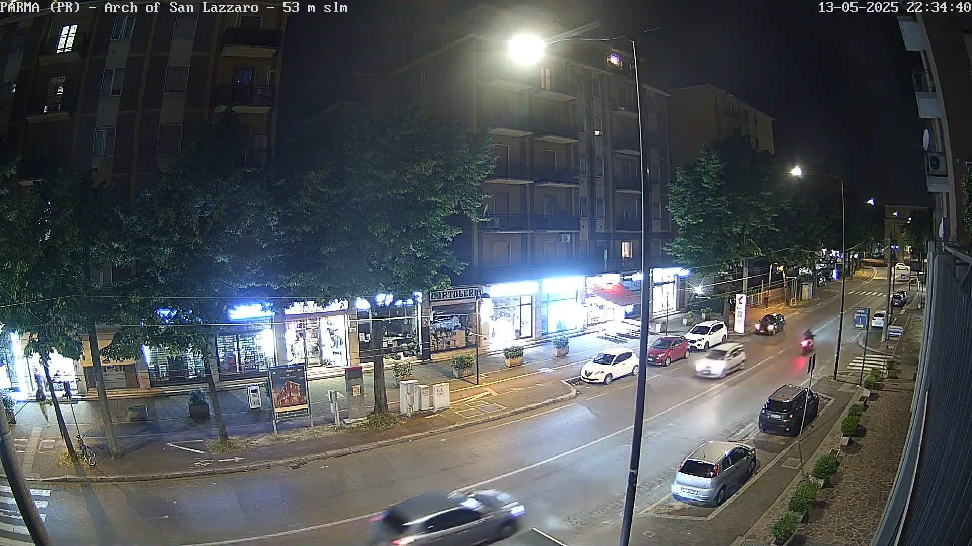 A nighttime street scene shows several cars driving on a road lined with shops and trees under streetlights.