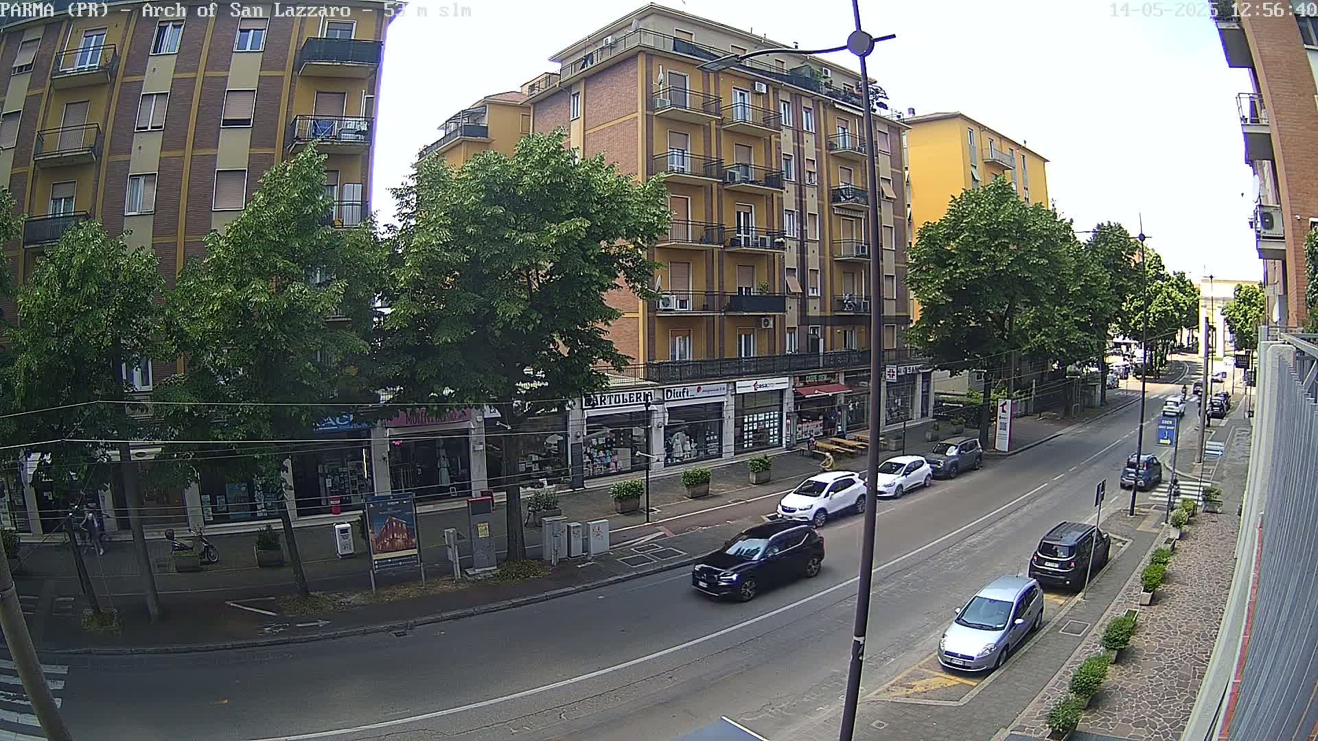 A sunny street scene shows a multi-story yellow building with shops on the ground floor, lined with trees and several cars parked along the curb.