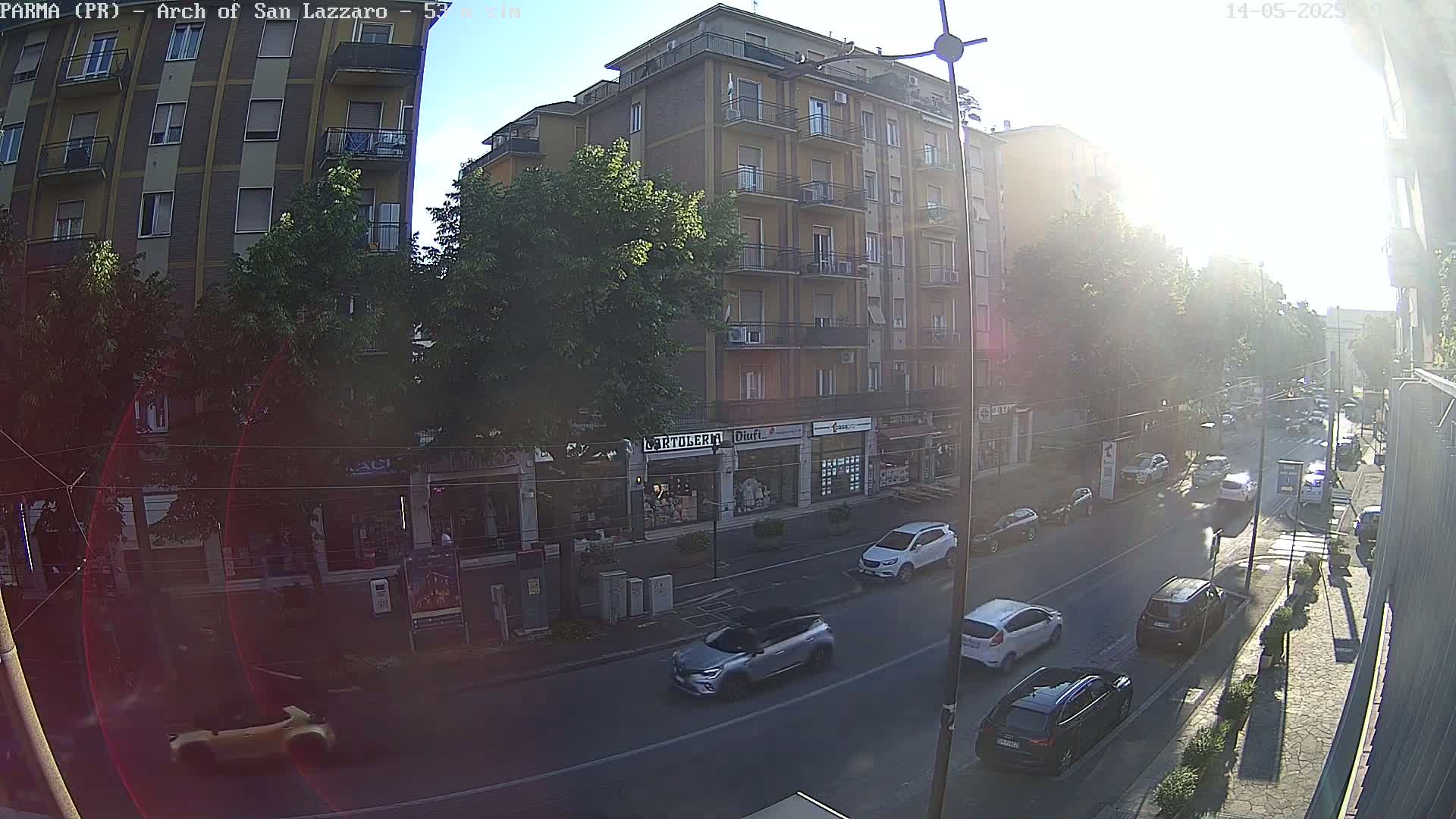 A sunny day reveals a city street lined with shops and a multi-story building, with several cars parked and driving along the road.