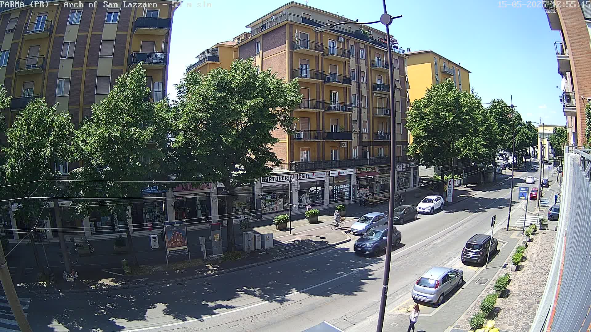 A sunny street scene shows a multi-story building with shops on the ground floor, several cars parked and driving along the road, and pedestrians walking on the sidewalk under leafy trees.