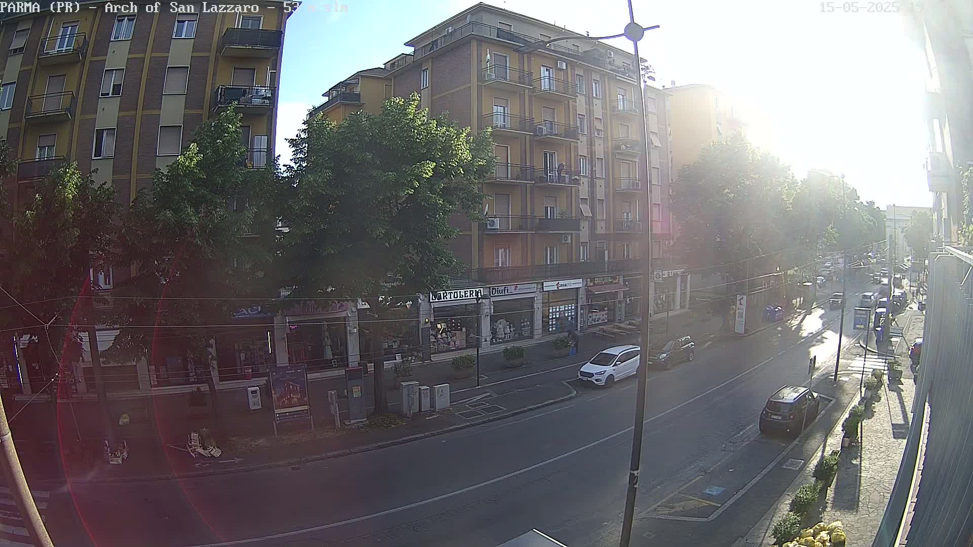A sunny street scene shows a multi-story building with shops on the ground floor, several cars parked and driving on the road in front, and trees lining the sidewalk.