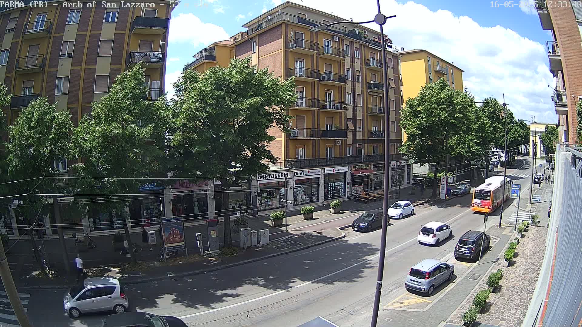 A sunny day shows a city street with cars parked and driving past multi-story buildings and shops, lined with trees.