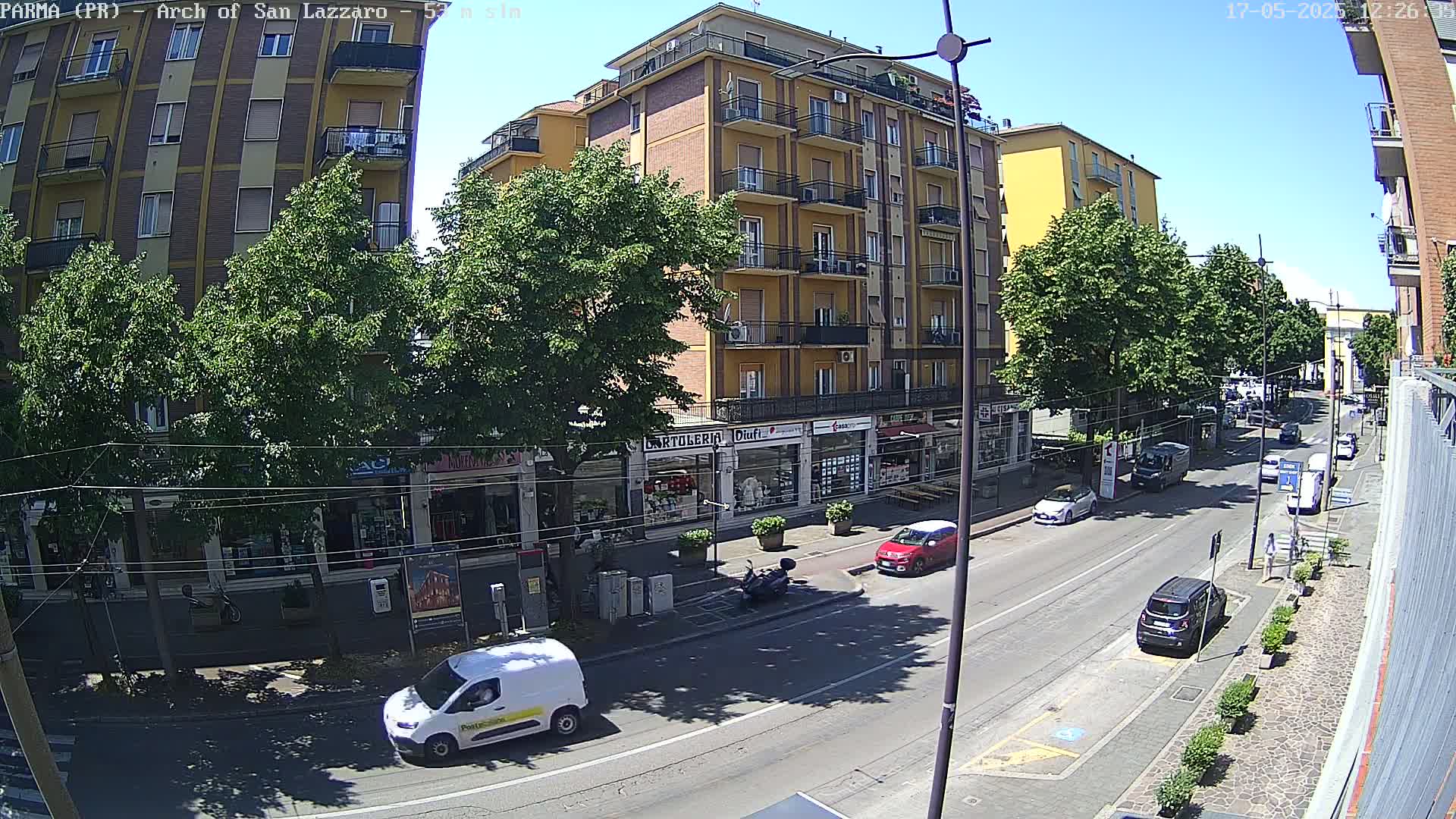 A sunny street scene shows a multi-story building with shops on the ground floor, lined with trees, and several cars and a van parked along the road.