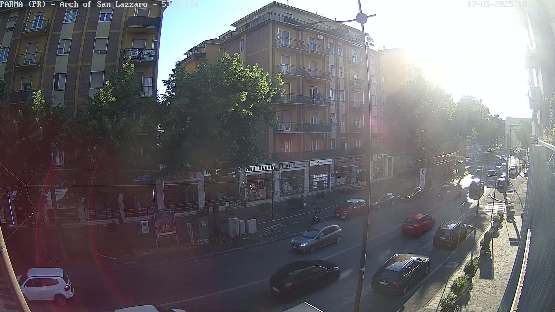 A sunny street scene shows a multi-story building with shops on the ground floor, several cars driving on the road in front of it, and trees lining the street.