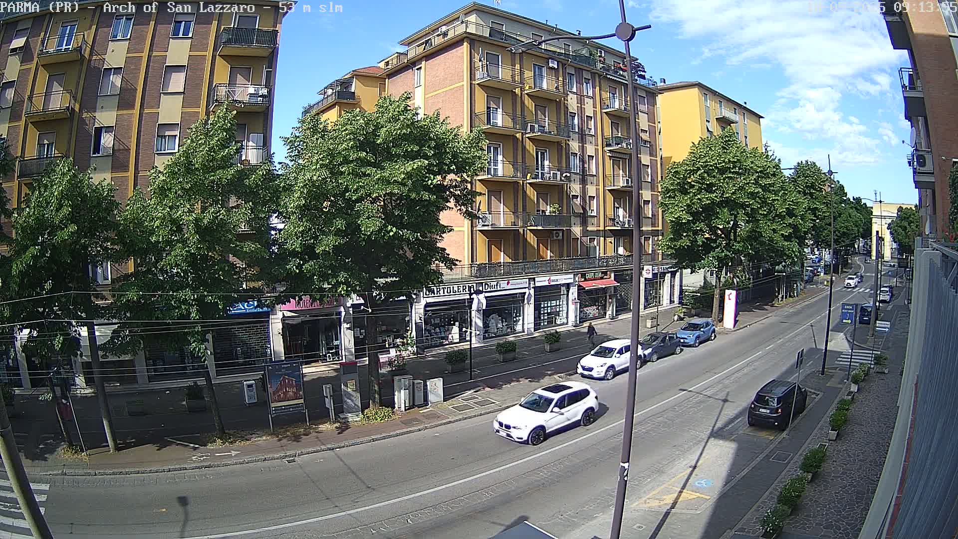 A sunny day reveals a city street scene with several cars parked and driving past multi-story buildings and shops, lined with trees.
