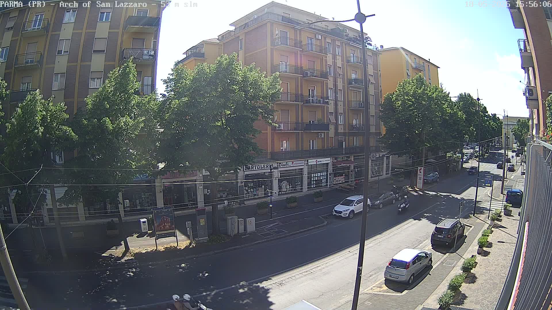 A sunny city street scene shows a multi-story building with shops on the ground floor, several cars parked along the street, and some pedestrians and motorcyclists.