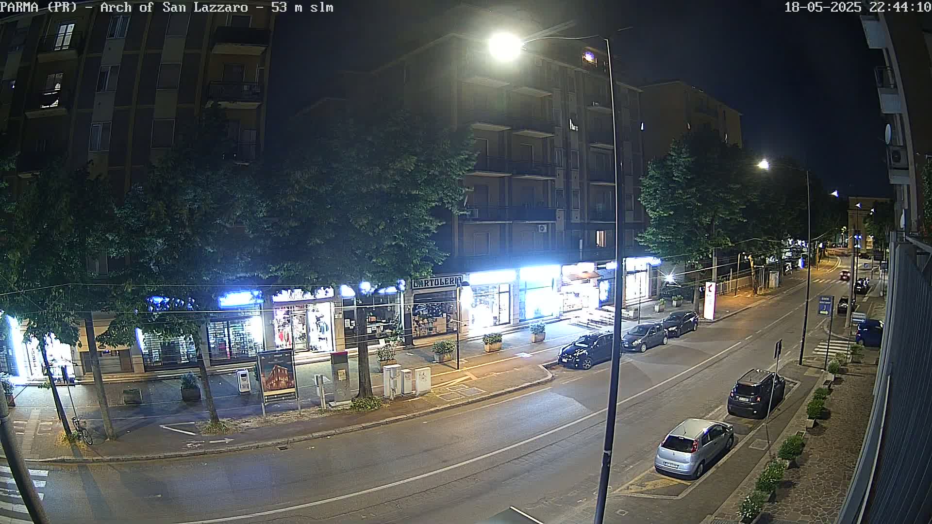 A nighttime street scene shows several cars parked along the curb in front of shops and buildings, under streetlights.